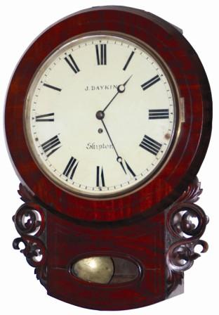 English short drop time only wall clock, certainly a cut above the usual English drop clocks we get. This one is mahogany veneered, 22" high, has some great carved side decorations, round wood bezel, four pegs to hold the top to the box, a bottom hinged door to access the pendulum, a hinged door on the side, and the oval glass at the bottom surrounded by rounded molding. — Antique Clocks Price Guide