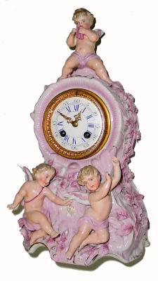 C.1890, German, Lenzkirch, Porcelain china clock with 3 dimensional leafy applique; musical themes, the winged cherub at top playing a flute, and a pair of cherubs frolicking below the dial. — Antique Clocks Price Guide