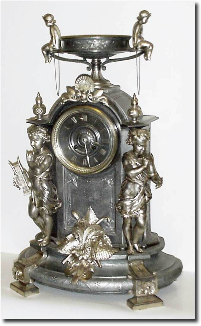C.1890, French, Silvered Spelter Figural clock, an unusual design with young boys and masks, one boy holding a harp, and the other a writing instrument; the case surmounted by a large urn with two young boys seated on the side handles. — Antique Clocks Price Guide