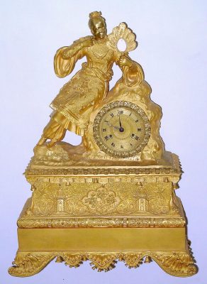 C.1840, French, Ormolu Figural Clock, Asian Motif, a lady clad in ceremonial dress with small pointy shoes, her braided hair rolled on top of her head, and with small blossoms in her hair, with a feather fan in her hand, the frieze below the lady with two pagodas or temples, each with a hanging bell, and little bells cast into the edge of the case all round the center and the base. — Antique Clocks Price Guide