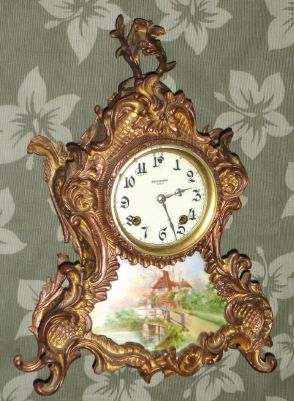 New Haven Clock Co., ROSEMONT, gilt spelter mantel clock, with a shaped plaque showing a boy fishing in front of a country cottage, girl watching, 8-day time and strike. C.1913. — Antique Clocks Price Guide