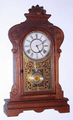Waterbury Clock Co., MELROSE, walnut gingerbread clock, with 8-day time and gong strike spring movement, and nickeled pendulum with glass center. C.1881. — Antique Clocks Price Guide