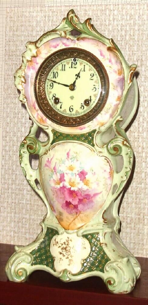 Ansonia Clock Co., Royal Bonn, "No. 506", slender porcelain clock in the French style, with one piece porcelain dial, creamy colored with red accents between the numerals; the white case with misty green edges and dark green accents + gold highlights, and a large cameo with floral spray below the dial. Sides of case decorated with violet vines. — Antique Clocks Price Guide