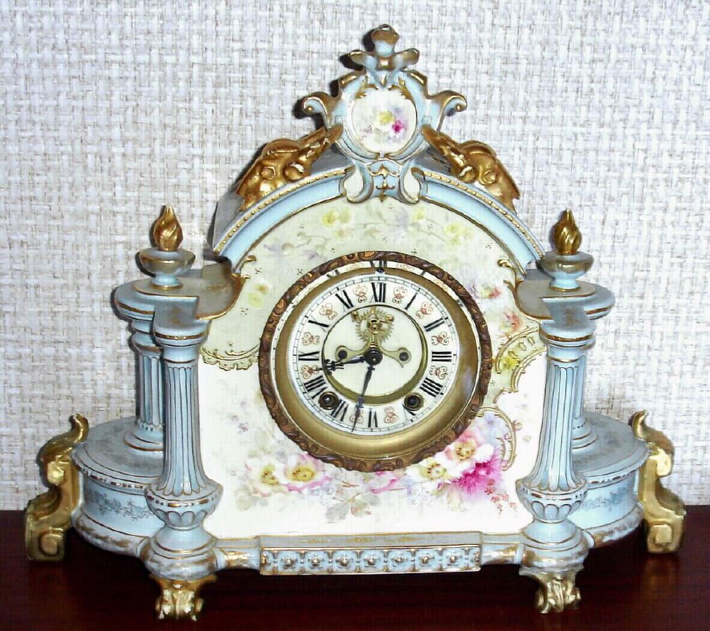 Ansonia Clock Co., Royal Bonn, "LA CHAMPAGNE", porcelain clock in the French style, with open escapement on the dial center, 8-day gong striking. The extra wide case of white china with light blue "roof", sides and accents and with pastel flowers. Profuse gold highlights, including the animal skulls that flank the top crest. — Antique Clocks Price Guide