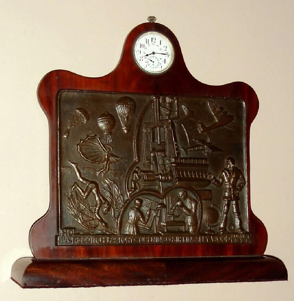 Desk clock on walnut stand with quality Swiss fully jeweled stem wind pendant set one day watch movement with 24 hour Arabic numeral porcelain dial set in scrolled top above bronze plaque in Diego Rivera style depicting factory scenes commemorating the opening of the H.D.A. factory in Redditch, England in 1939, c1940 — Antique Clocks Price Guide