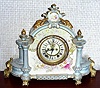 Ansonia Clock Co., Royal Bonn, "LA CHAMPAGNE", porcelain clock in the French style, with open escapement on the dial center, 8-day gong striking. The extra wide case of white china with light blue "roof", sides and accents and with pastel flowers. Profuse gold highlights, including the animal skulls that flank the top crest.