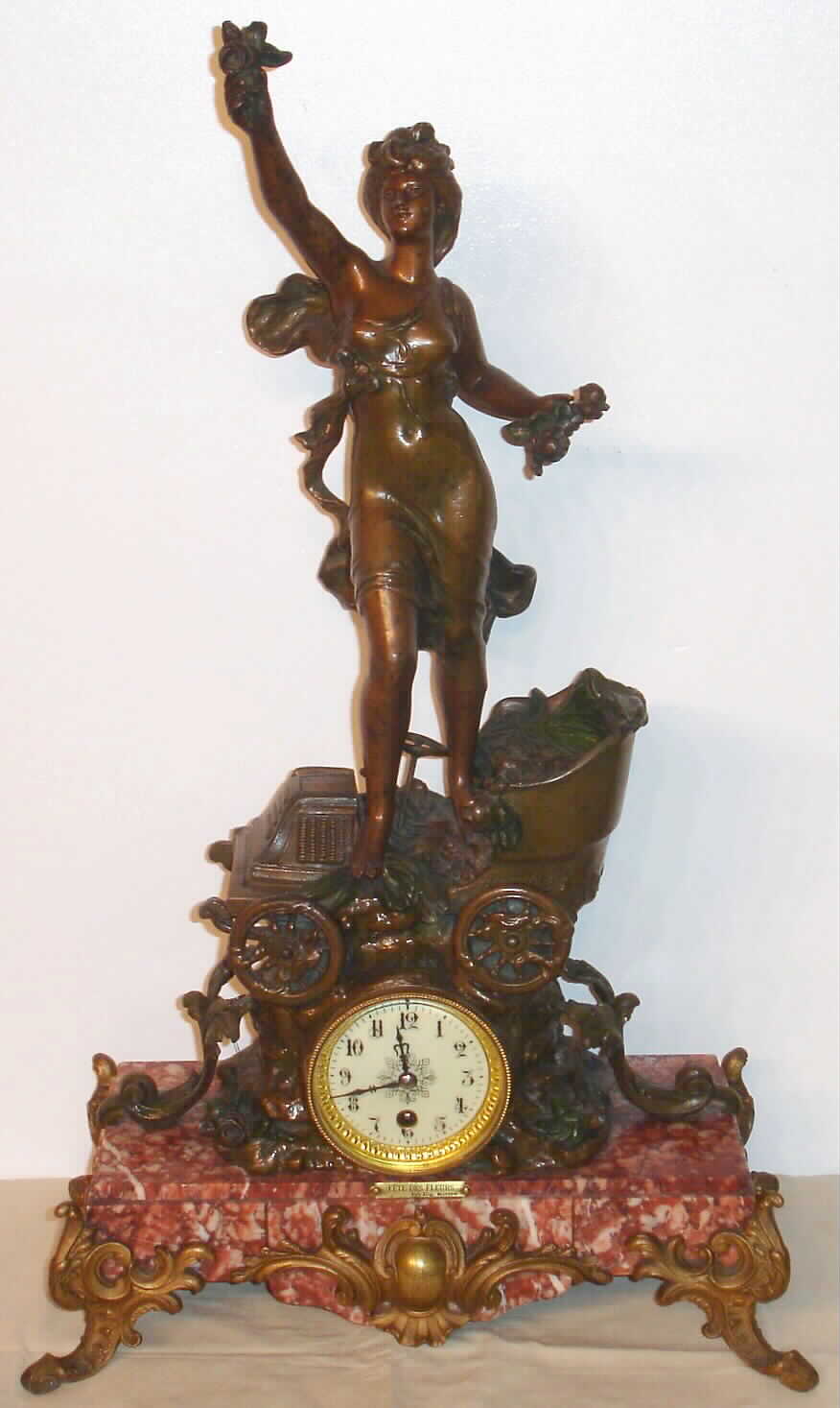 French, patinated spelter and marble figural clock of a "Car", a stylized version of a C.1900 motor car with steering wheel, with a statue of a flower girl standing on the car, the car mounted to a large rectangle of red and pink variegated marble, and the original title plaque "Fete de Fleurs". With a quality 8-day movement and porcelain dial. — Antique Clocks Price Guide