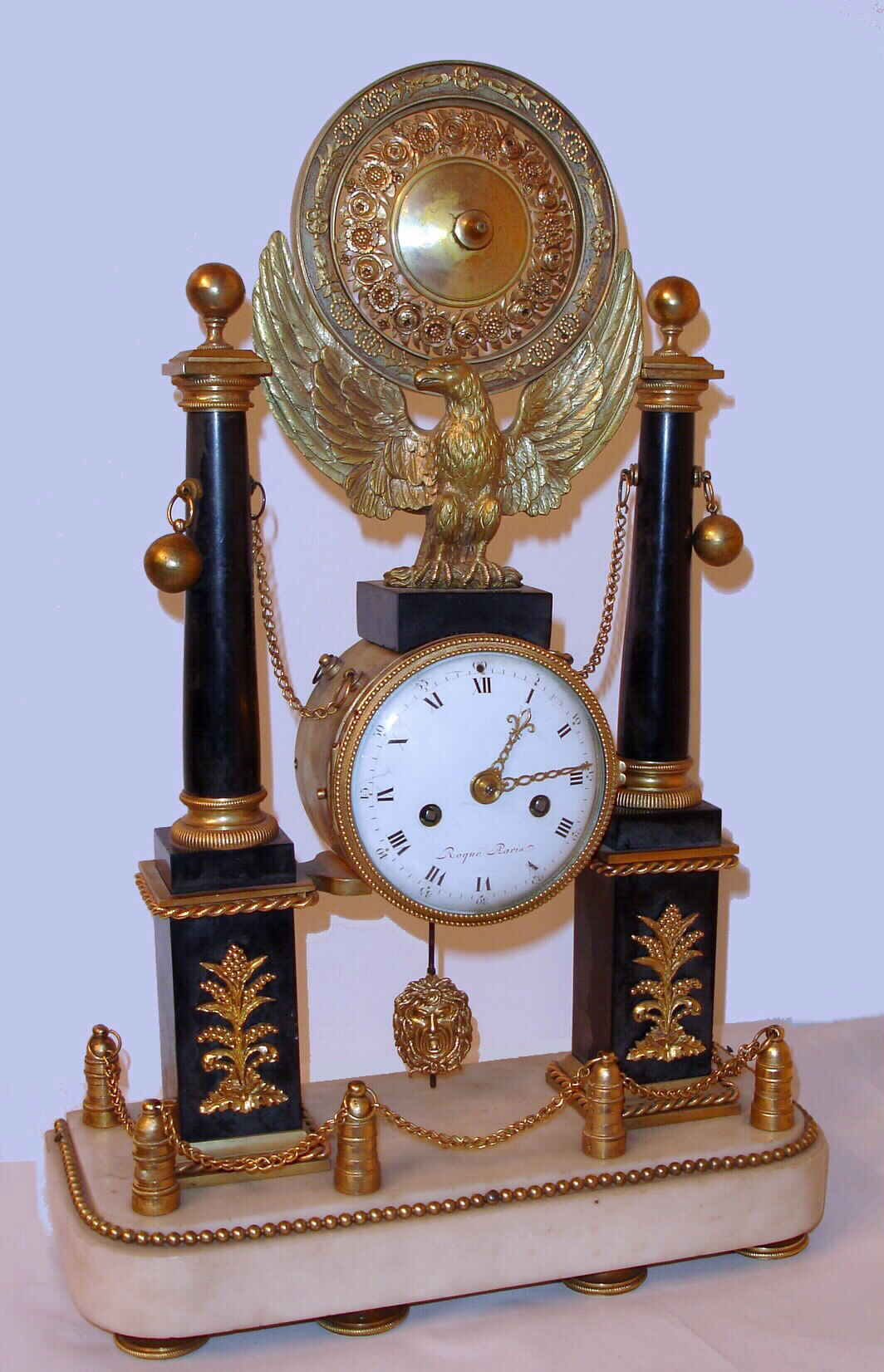 Roque, Paris, French architectural mantel clock with 8 day time and strike on bell spring driven movement engraved with the maker's name and No. 1282. The clock rests on white marble base supporting six posts linked by chain with two marble pillars with ormolu appliques and finials centered by brass spread wing eagle perched above movement supporting ornately floral embossed medallion. The porcelain dial has intricately pierced brass hands and is signed Roque Paris (fired in), c1820 — Antique Clocks Price Guide