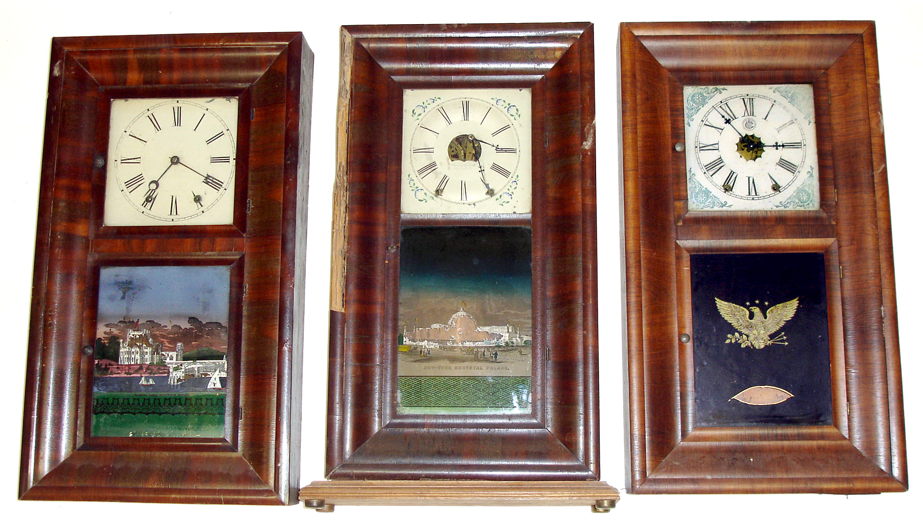 Three 8 day ogee Connecticut weight driven shelf clocks- the first is a Chauncey Goodrich dial, later added base, and old New York Crystal Palace tablet (and prior insect damage visible behind loose veneer strip), a Jerome and Co. 2 door with old English castle tablet, and a Waterbury 2-door with bold patriotic tablet. All 30" tall — Antique Clocks Price Guide