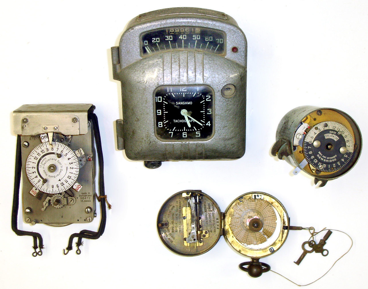 Four metal case specialty timekeepers. Oldest is C.1880 portable pocket watch watchman's clock, made in Germany. Sangamo "Tachograph" is automotive clock, speedometer and odometer, and is the largest at 8" tall. Swiss 13 jewel electric time switch is from Sauter, Basle. New Bridge with British Patent is cylindrical time switch with unusual month indicator. — Antique Clocks Price Guide