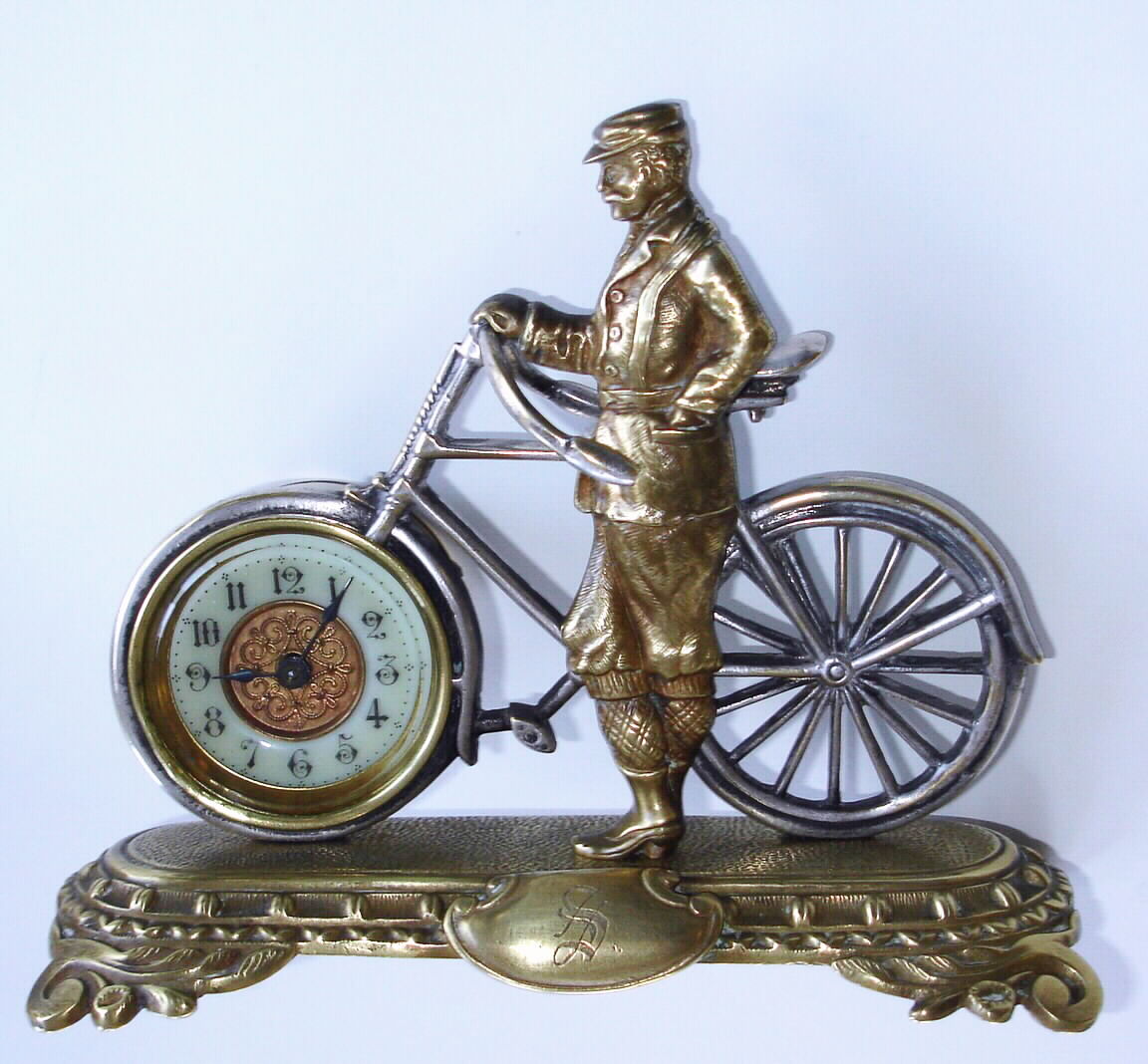 English, British United Clock Co, Ltd, Birmingham, a cast brass bicycle and male rider, in period riding clothes, and with a 30 hour rear wind and set movement in place of the front wheel, the whole on a shaped brass base with a cartouche bearing the initial "S", c1910 — Antique Clocks Price Guide