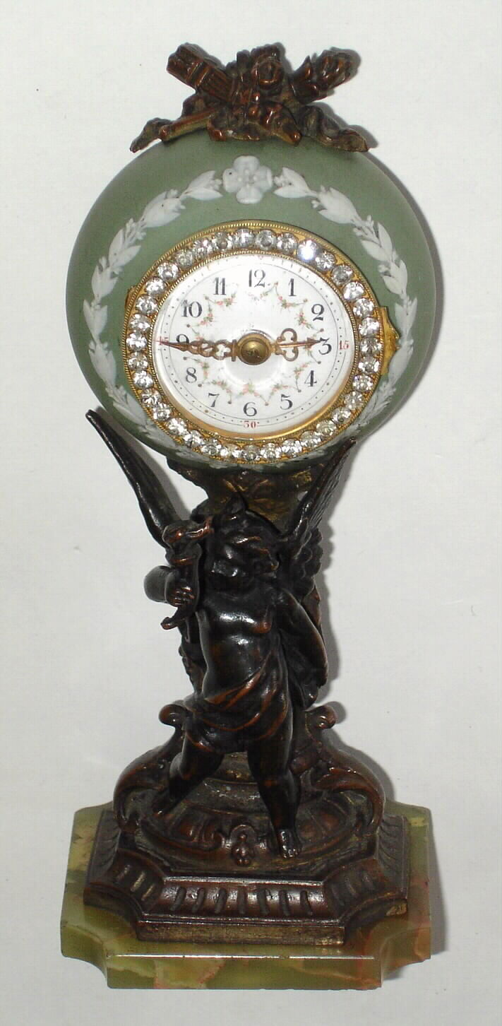 French, miniature timepiece in a light green Wedgewood case, resting on a patinated spelter stand (pole with a torch bearing winged man), the whole resting on a green onyx base. A small round French movement has a tiny lever platform [yes], in good order, c1900 — Antique Clocks Price Guide