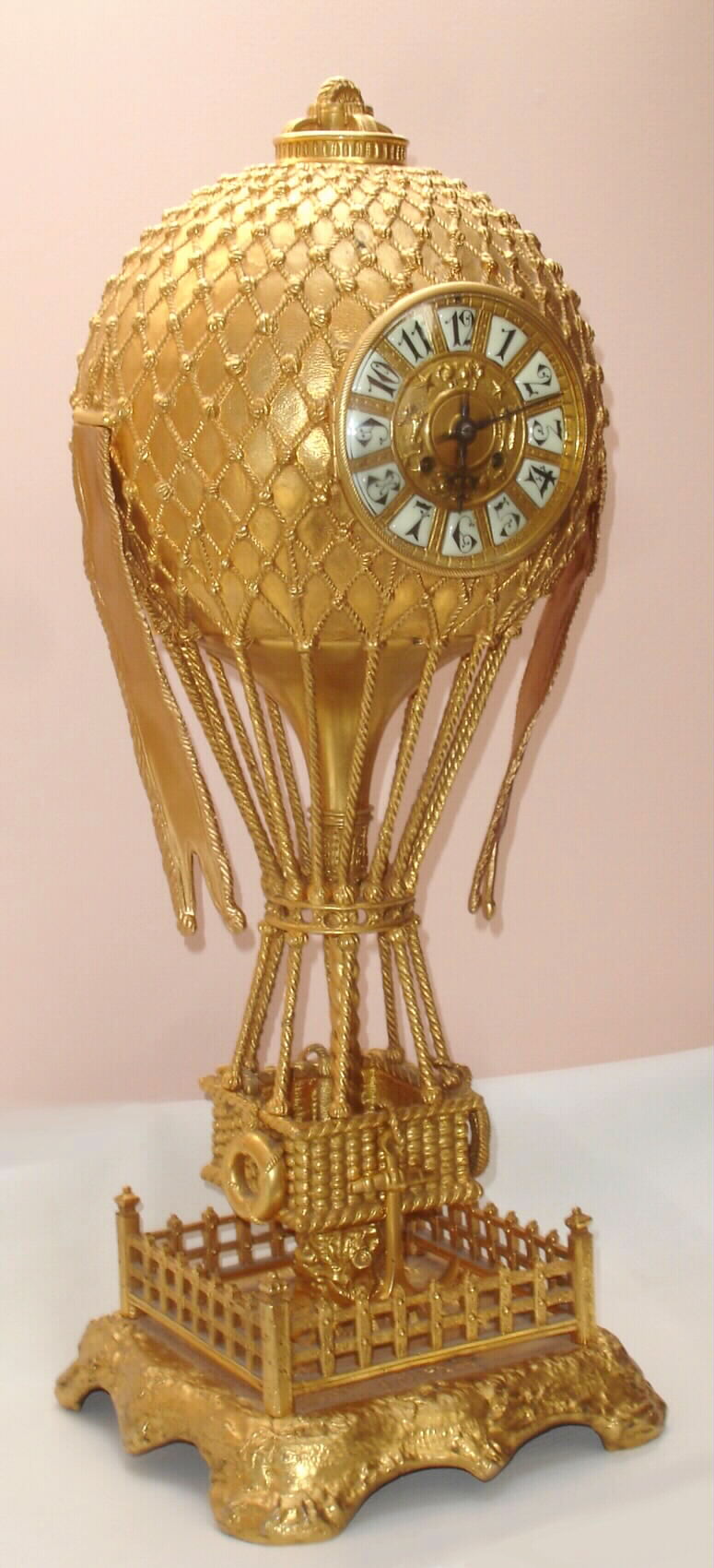 French, Large Cast and gilt bronze Balloon Clock, the case depicting a hot air balloon with a woven basket below, and is anchored to the ground within a fenced platform. Has a gold dore finish. Dial has applied Arabic numerals and fancy black hands; typical round French brass time and strike movement, signed Japy Freres, c1890 — Antique Clocks Price Guide