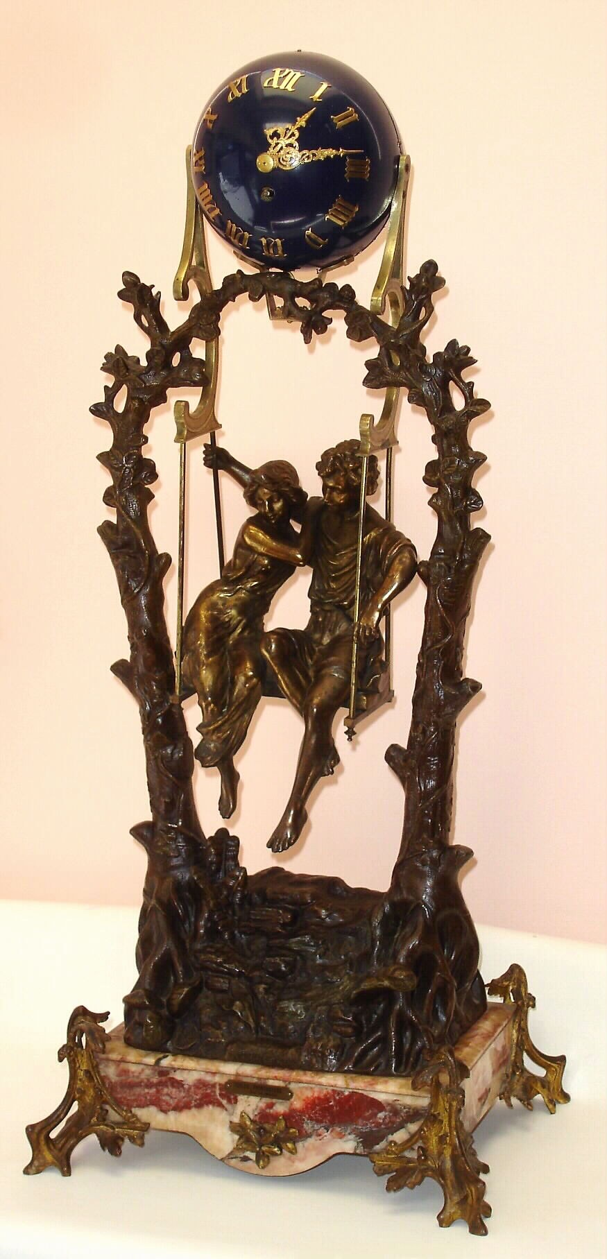 French, very large swing clock, Patinated spelter tree limbs entwined at the top to form the support for the swinging clock. A romantic couple are seated in a swing, appearing to suspend by rope, The original marble base with a brass title plaque: "Idylle Printaniere, par Geo Maxim", the couple swinging from side to side without apparent impulse, c1890 — Antique Clocks Price Guide