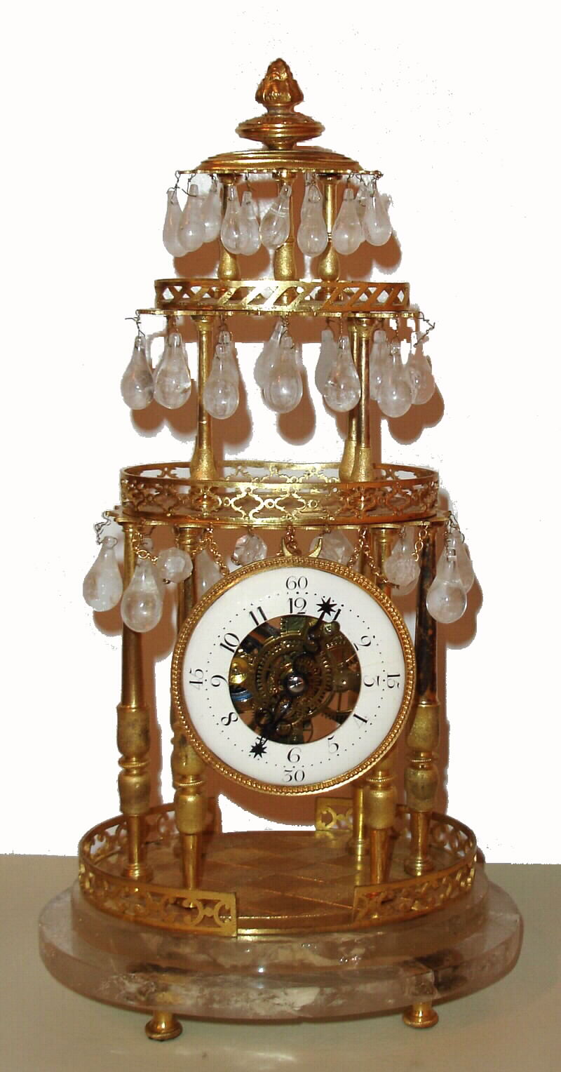 French mantel clock, 8 day, time only, spring driven, verge fusee movement and balance wheel in a three tiered gilded brass pillared case with canopy top resting on a rock crystal with four gilded feet. Tear drop rock crystal prisms are suspended from the three tiers. The white enamel chapter ring exposes the skeletonized movement, c1860 — Antique Clocks Price Guide