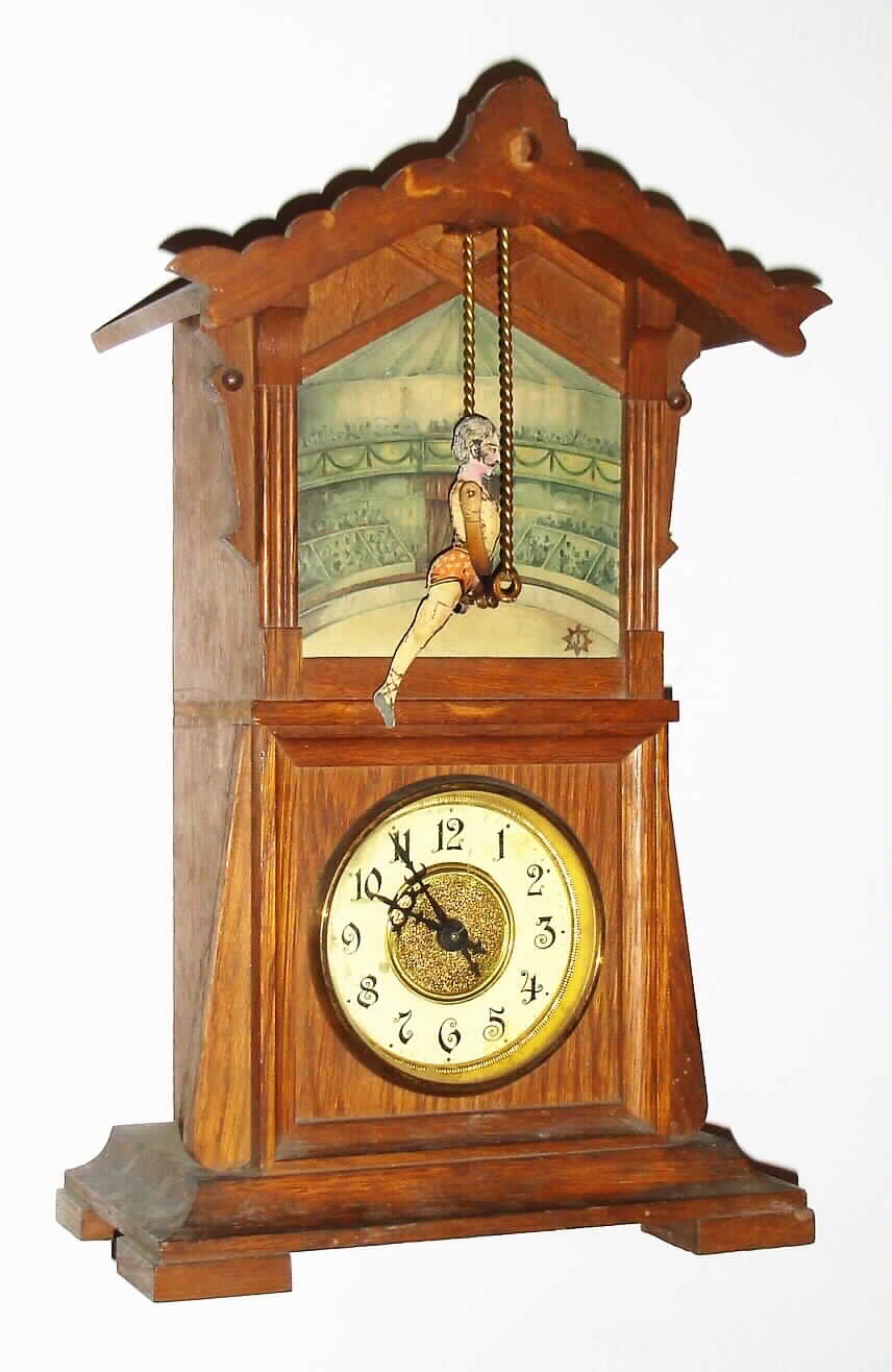 Junghans, Germany, "Acrobat" model novelty, with automated figure, spring powered thirty hour two train movement, one train for time, the other for the performer, oak case with original finish, acrobat on trapeze in front of lithograph scene of the interior of a circus tent with spectators, Junghans logo at lower right, coated paper dial with Arabic figures, dial trim and center in polished, lacquered brass, blued steel hands, — Antique Clocks Price Guide