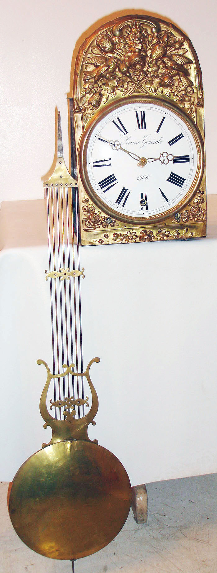 French 3 train Morbier clock, the large movement with recoil escapement, and quarter striking on 3 bells, striking the hour on a large bell, large gridiron style lyre pendulum with large convex brass shell ball, driven by three gold painted cast iron weights, white Roman numeral enamel dial marked "Procure Generale 1906", pierced brass hands,  the brass surround stamped in high relief and depicting two birds, leaves and flowers, decorated sheet iron doors on either side of the case provide access to the movement, and sheet iron back stamped with concentric squares and circles, c1906 — Antique Clocks Price Guide