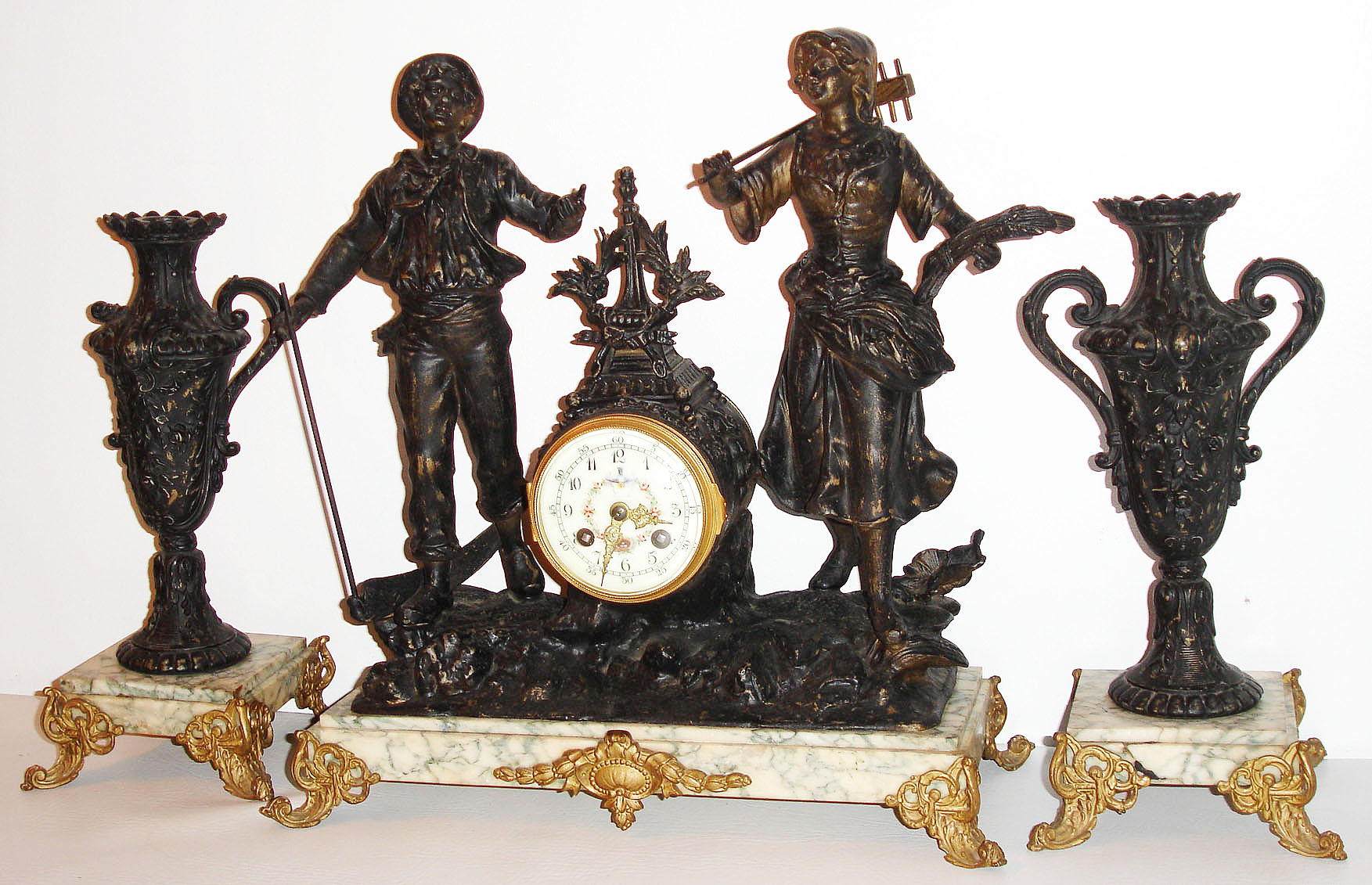 France, three piece mantel garniture, 8 day, time and strike, pendule a Paris movement, the clock case with a harvest theme, male and female figures with rake and scythe, clock case decorated with flower garlands, white enamel Roman numeral dial with a polychrome enamel center featuring a bird holding floral garlands supporting a nest with eggs above the 6, gilt Louis XV style hands, gilt bezel with convex beveled glass, all on a white marble base with blue green veining, supported by scrolled feet, the urn form candle holders with handles and floral ornament, set on bases matching the clock, c1880 — Antique Clocks Price Guide