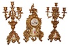France, 3 piece mantel garniture in gilt brass, the clock and candelabra in the rococo style, heavily laden with foliage and C scrolls, the case front with inset polychrome enamel decorated porcelain panel featuring a man and woman in fancy dress, Roman numeral porcelain dial with gilt chapter and cupid at the center, the gilt candelabra similarly ornamented, but each with a single figure, and 8 days, time and strike, pendule a Paris movement by Japy Freres, c1910