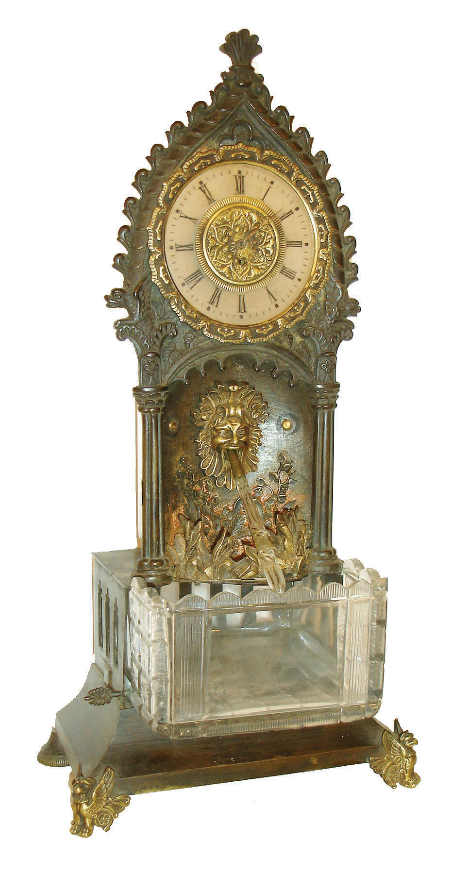 France, an unusual table clock with fountain, patinated bronze case in the Gothic taste, the base containing a cut a cut glass reservoir, the front corners with gryphon feet, case front with niche, trimmed with cast foliate ornament and grotesque mask, a twisted glass rod which rotates under power of a concealed movement simulates water pouring from the mouth of the mask, frosted glass dial with painted Roman numerals, 30 hour Swiss verge movement, c1850 — Antique Clocks Price Guide