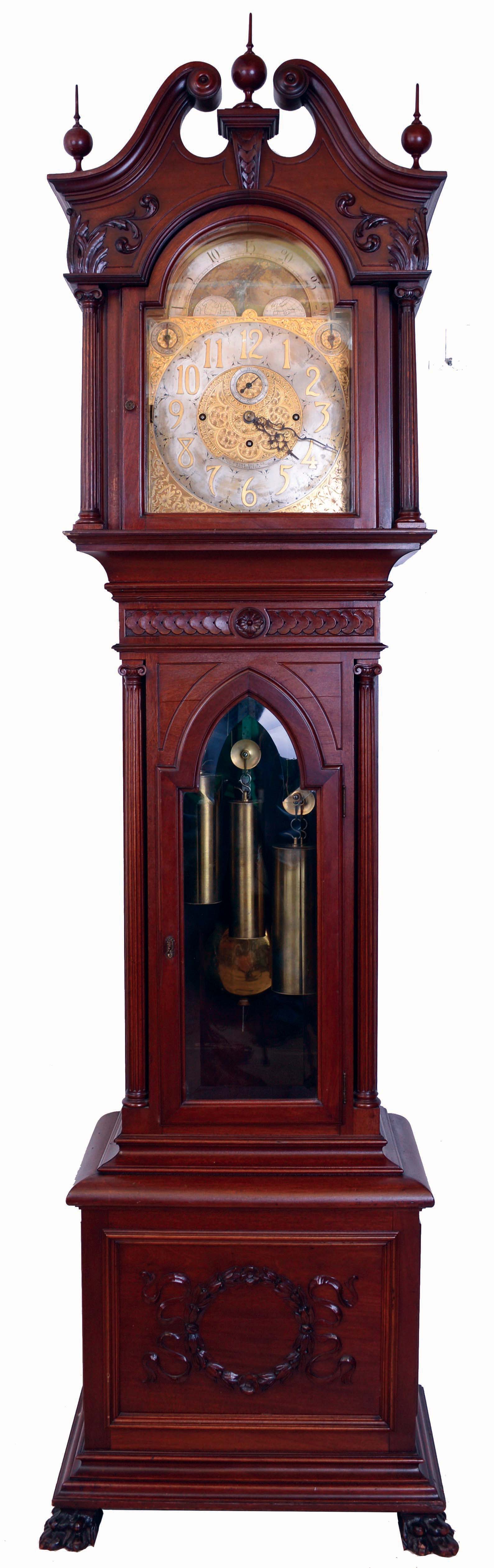 John J. Elliott, London, England (attribution) for Simons Bros. Co., Philadelphia, Penn., tall clock, time, strike and Westminster and Whittington chime on 8 bells and four gongs, 8 days, three brass weight driven movement in a mahogany case with full reeded Corinthian columns, beveled Gothic arch glass in trunk door, paw feet, carved wreath on center of plinth and carving above and below trunk, The hood has broken arch top with turned rosettes, fretwork sides, carved leaves on chimney under center finial and full reeded Corinthian columns. The brass dial has ornately cutout and engraved brass corner spandrels and profusely engraved dial center, silvered chapter ring with applied brass numerals, hour strike / silent and Westminster and Whittington selectors and moon phase dial, c1910 — Antique Clocks Price Guide