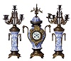 French, Blue and White, Delft style mantel garniture set [3 piece], of brass and china, the candle stands with bronze, winged gargoyles supporting each candle, and with fancy porcelain dial with hand painted cherub below 12
