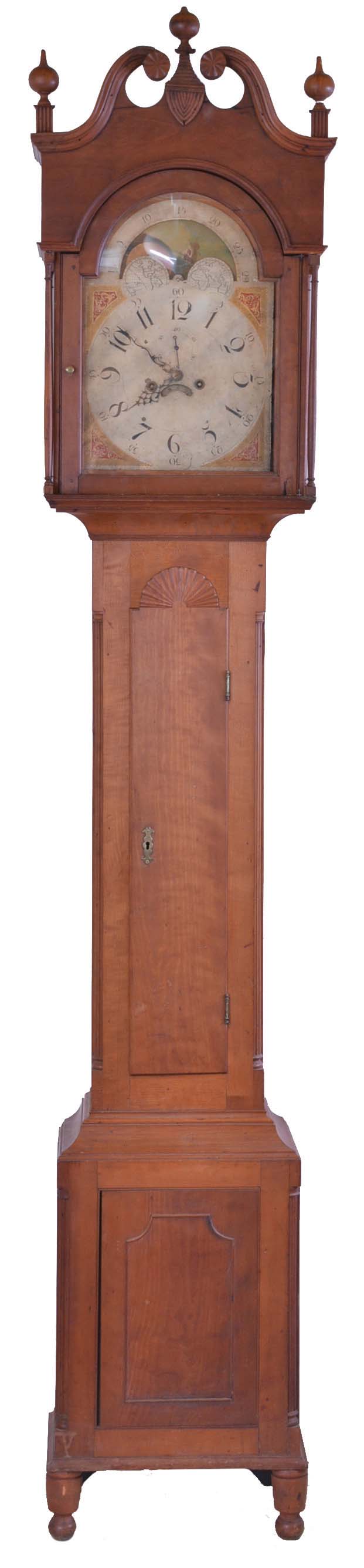 Pennsylvania, tall clock, 8 day, time and strike, weight driven movement in a cherry case with broken arch set with carved rosettes, carved plinths with three wooden finials, slender turned columns, sunburst carving at top of trunk door and reeded corner columns on trunk resting on a base with beaded center panel and turned front feet and rear bracket feet. The unsigned painted metal dial is attributed to William Jones of Philadelphia and includes moon phase with ship flying American flag. — Antique Clocks Price Guide