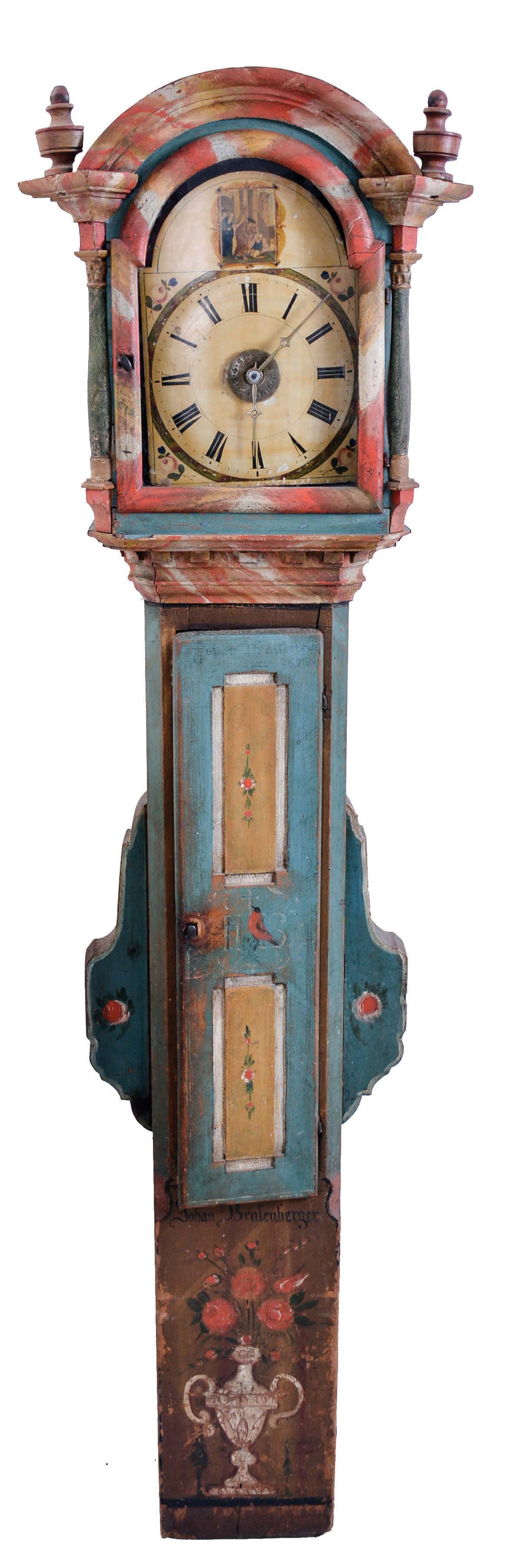 Germany, dwarf tall clock, 30 hour, time, strike and alarm weight driven wooden plate movement, signed on the reverse in old German script, in a paint decorated case, signed "Johan Bratenberger" over a vase full of flowers with undecipherable initials on trunk door, is decorated with a robin, and floral decorations on either side of hood. The case has decorated wings to accommodate pendulum swing. The paint decorated wooden dial has a religious scene in arch. — Antique Clocks Price Guide