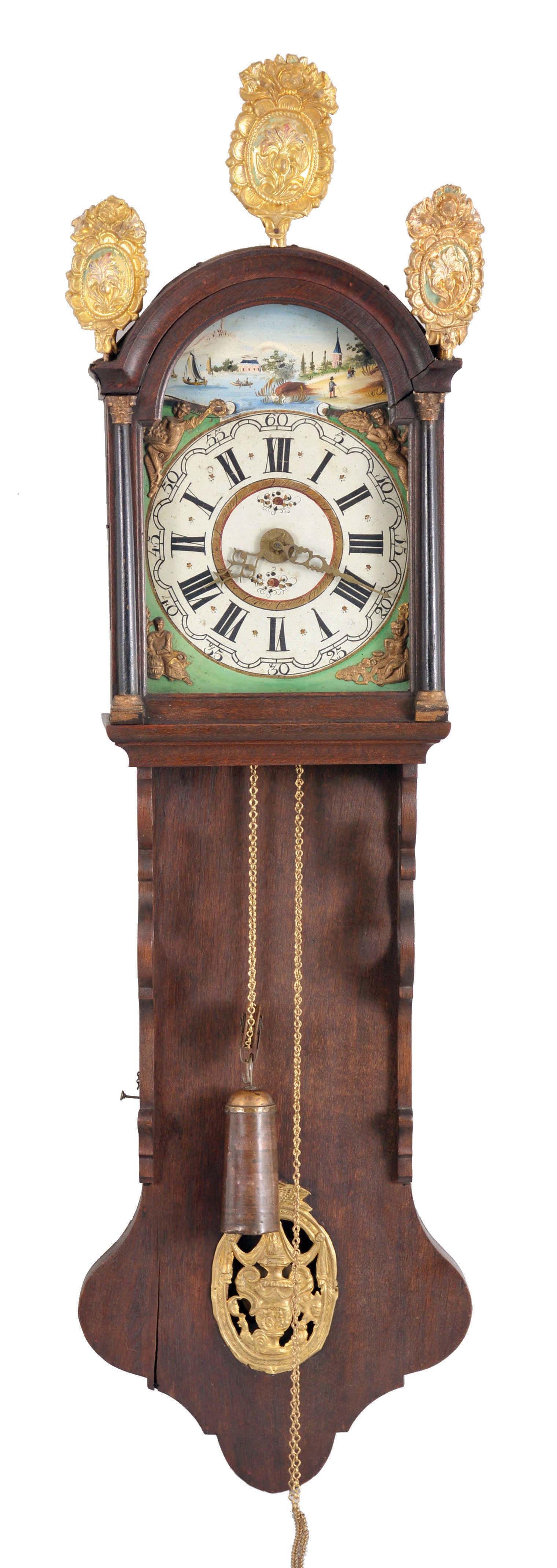 Holland, Staartklok, oak case with arch top bonnet, the door with half columns, gilt capitals and bases, and fancy stamped brass finials, the pendulum aperture with stamped brass ornament, arcaded, Roman numeral painted dial with polychrome landscape shoreline scene in the arch, and with applied, stamped brass figures at the corners, cast brass hands, thirty hour time and strike movement with alarm, tapered, copper clad weight — Antique Clocks Price Guide