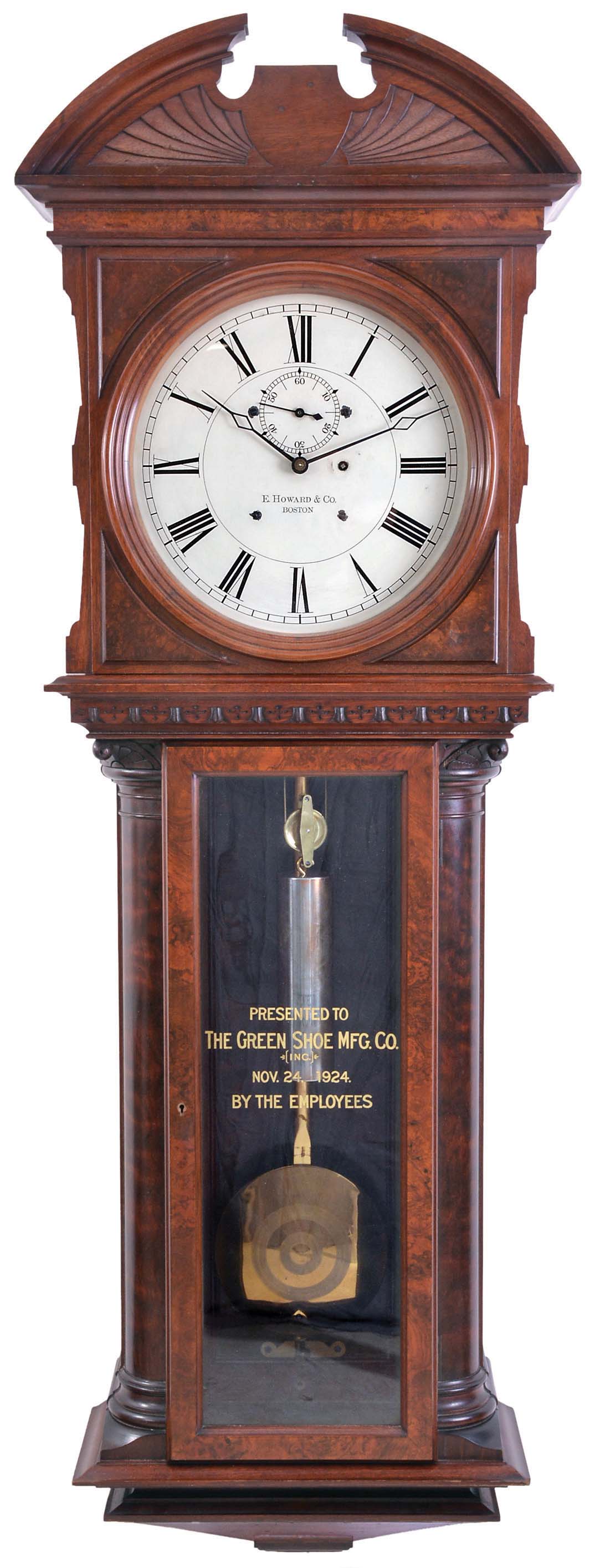 E. Howard &amp; Co., Boston, Mass., "Regulator No. 57", jeweler's regulator wall clock, the walnut and burl walnut case with bold quarter columns, having turned and carved capitals and turned bases, flanking the burl veneer trunk door with reverse painted and gilt presentation, "THE GREEN SHOE MFG. CO. (INC.) NOV. 24, 1924. BY THE EMPLOYEES", over a burl veneered, stepped, tapered bottom bracket, upper door with raised wooden spandrels enframing the dial, over a nicely detailed carved molding above trunk door, and fan carved, broken arch top, Roman numeral painted dial signed "E. HOWARD &amp; CO. BOSTON", 8 day, time only, weight driven movement with maintaining power and large brass, decorated bob and gold leafed pendulum rod, with cylindrical weight. — Antique Clocks Price Guide