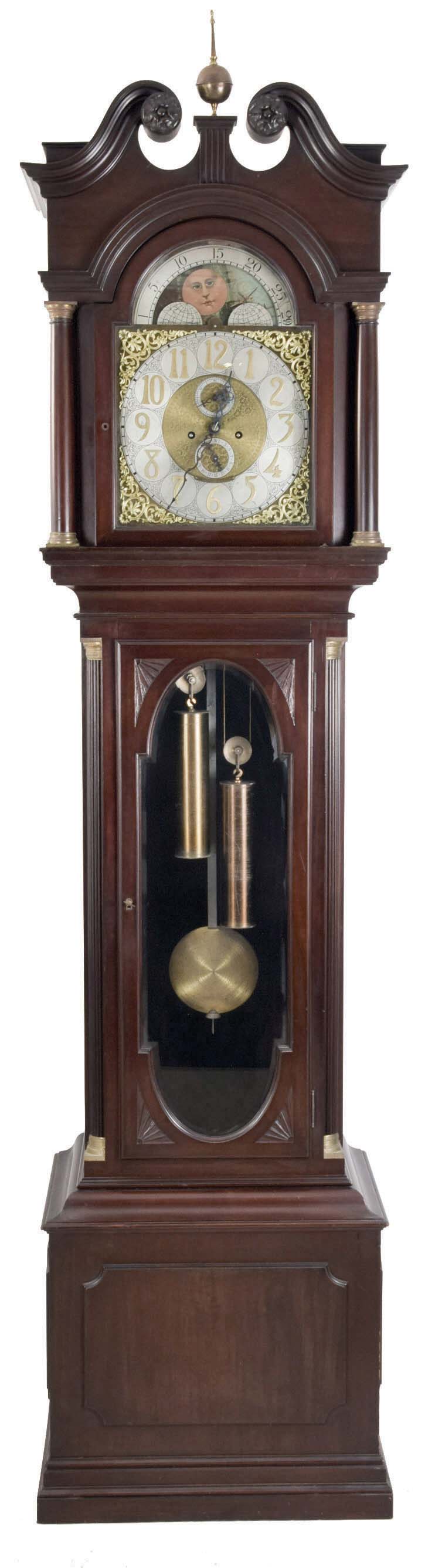Hall clock, 8 day, time and strike on gong, two weight driven movement by Elite (Germany) in a mahogany case with reeded corner columns on trunk with brass capital flanking beveled glass door. The hood has full masculine columns and carved rosettes on broken arch top. The brass dial has silvered chapter ring, applied brass numerals and spandrels, moon phase and silvered calendar and second's bit. — Antique Clocks Price Guide