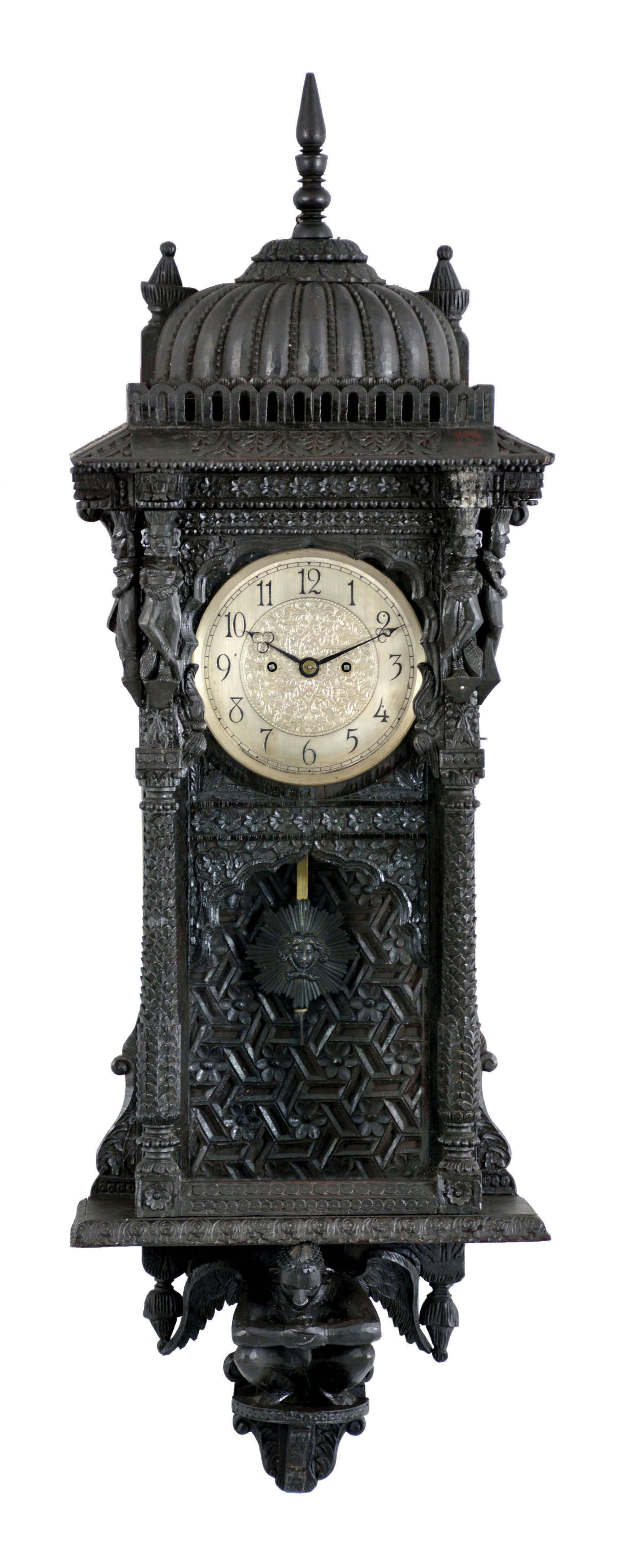 Wall clock, the case of tropical hardwoods, replete with Asian and Moorish inspired carving, the base with a winged figure, the door with leafy columns and with figures at the top corners, flat surfaces with geometric and naturalistic repeating motifs, the top with a ribbed dome, and turned finial, Arabic numeral silvered dial, blued steel trefoil hands, English 8 day, time and strike fusee movement, and pendulum with patinated Helios mask pendulum — Antique Clocks Price Guide