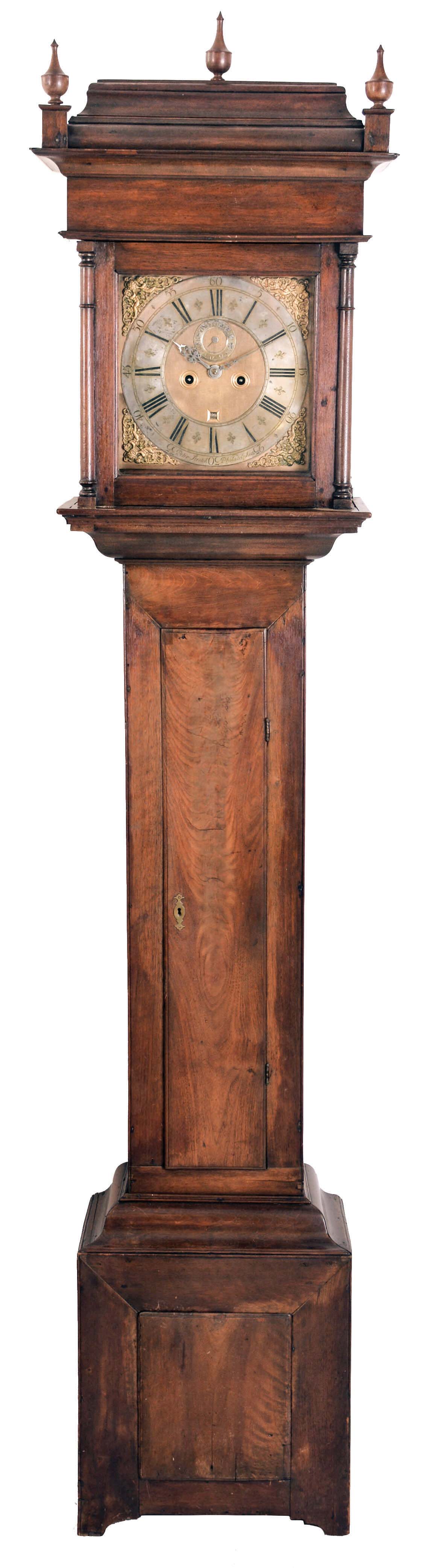 Peter Stretch, Philadelphia, 18th century tall clock, walnut case with caddy top above a deep, cavetto molded cornice, bonnet door with attached, turned columns, trunk with thumb molded, figured walnut door, and heavy ogee moldings at top and bottom, and the base with removable panel, feet cut from the front and sides, the square, composite dial with gilt, matted center, decorated winding holes, and Roman numeral, silvered chapter ring, pierced steel hands, 8 day, weight driven time and strike movement with count wheel between the plates, brass pendulum with wooden rod, and cylindrical lead weights — Antique Clocks Price Guide