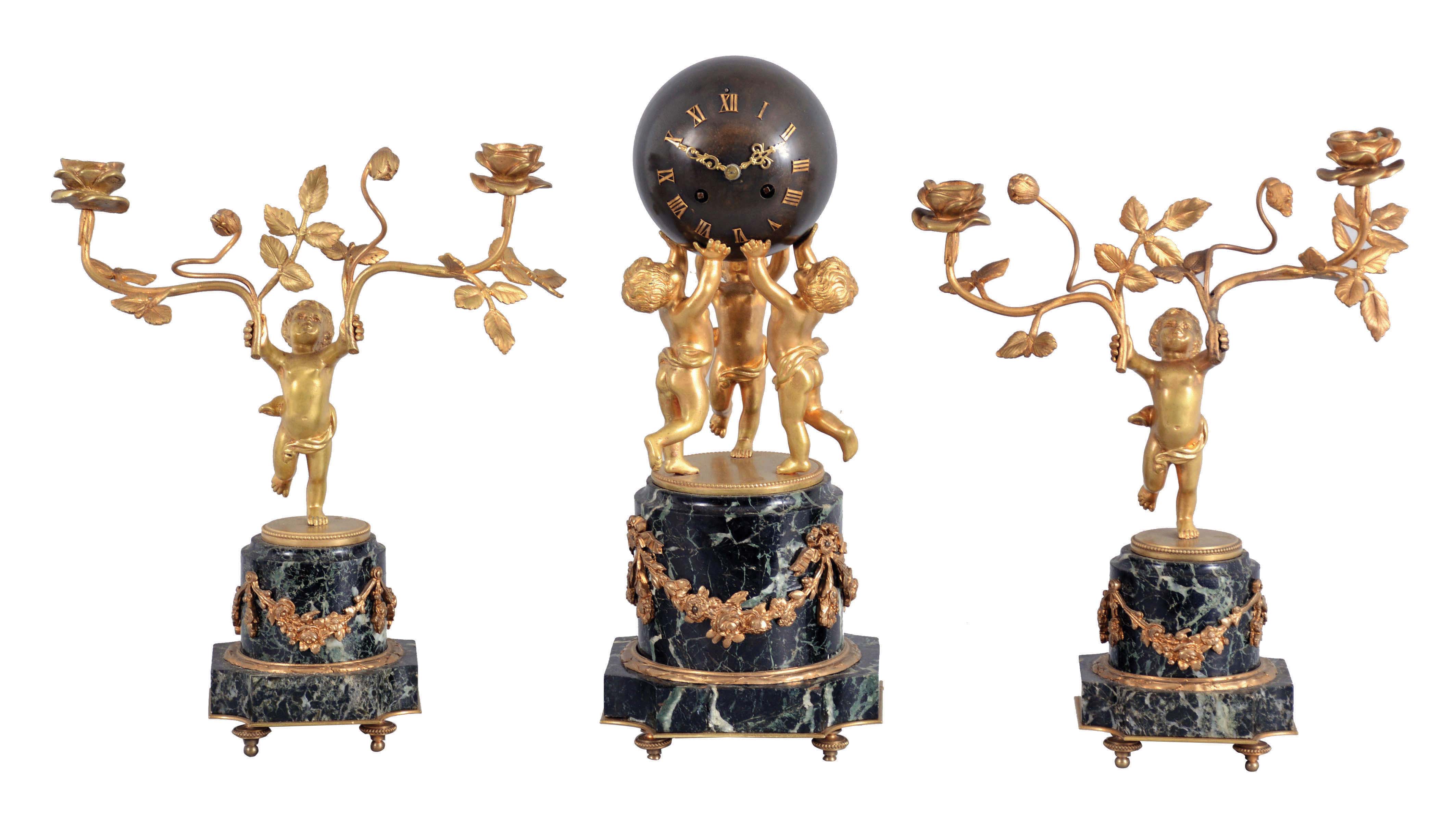 France, a three piece marble and gilt bronze mantel garniture, clock and candelabra on matching, variegated, green marble bases, the square plinths with incurved corners supporting attenuated columns draped with floral festoons, the larger, central base with three putti, holding aloft a patinated, spherical clock case with applied Roman numerals, and containing an 8 day, time and strike, pendule de Paris movement, the side pieces each with a single putto, with branching rose stem in each hand, the candle sockets of open rose form, clock dimensions listed — Antique Clocks Price Guide