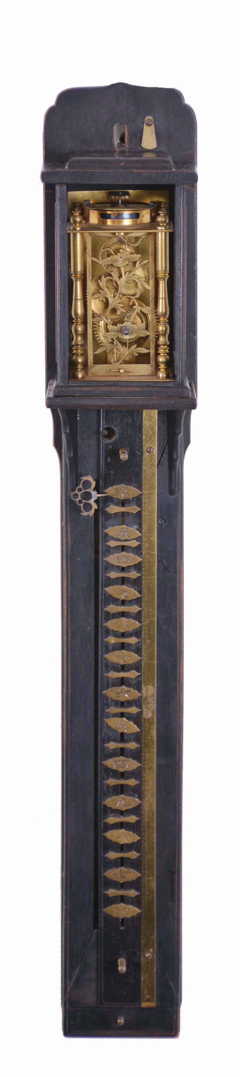 Japan, Edo period, striking wall clock, or Shaku-Dokei, commonly called "stick clock", the time movement of lantern style construction with crown wheel and verge driving the large, horizontal balance atop movement, delicately pierced front plate is intricately cut in the form of flowers, and is nicely engraved. The spring driven striking movement actually forms the weight that powers the time movement via a fine cord that winds around a horizontal barrel. Strikes on a bell. With movable numerals to compensate for the shortening days of winter. — Antique Clocks Price Guide