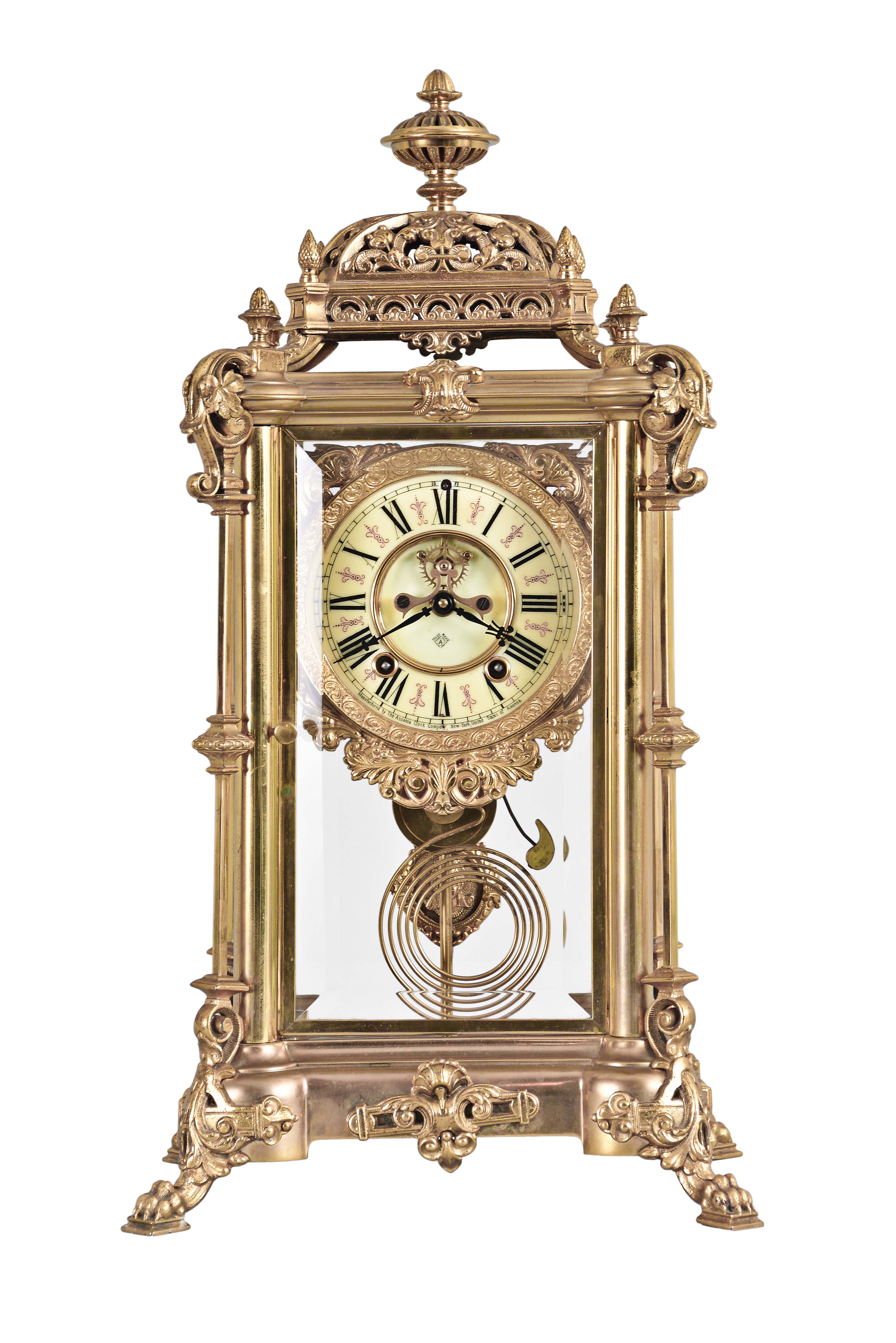Ansonia Clock Co., New York, "Excelsior" crystal regulator, 8 day, time and strike, spring driven movement with visible Brocot escapement in a gilt finished brass case on claw feet, cream porcelain dial with Roman numerals and red interstitial decorations, cast pendulum with female face, strikes on cathedral gong. — Antique Clocks Price Guide
