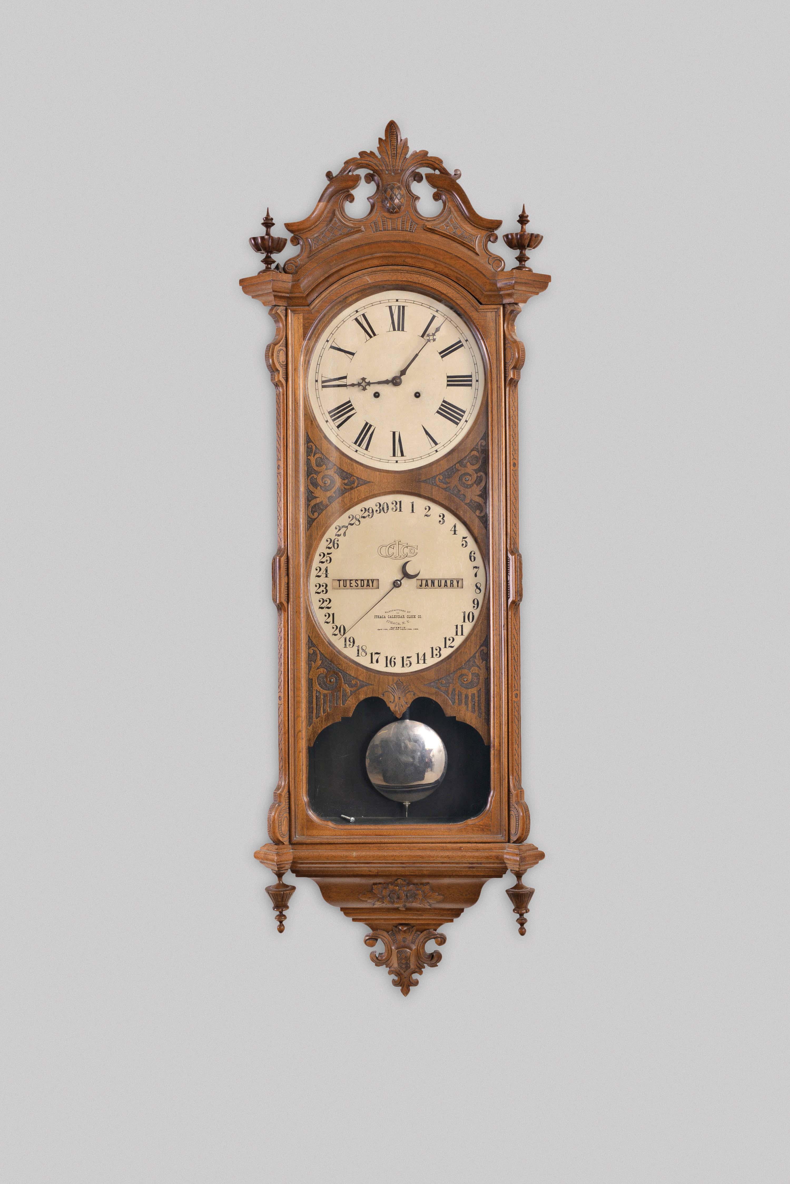 Ithaca Calendar Clock Co., Ithaca, New York, "No. 0 Bank", hanging 8 day, timepiece with perpetual calendar, weight driven movement in a walnut case with a carved pediment top, carved base and upper and lower finials with papered metal dials — Antique Clocks Price Guide
