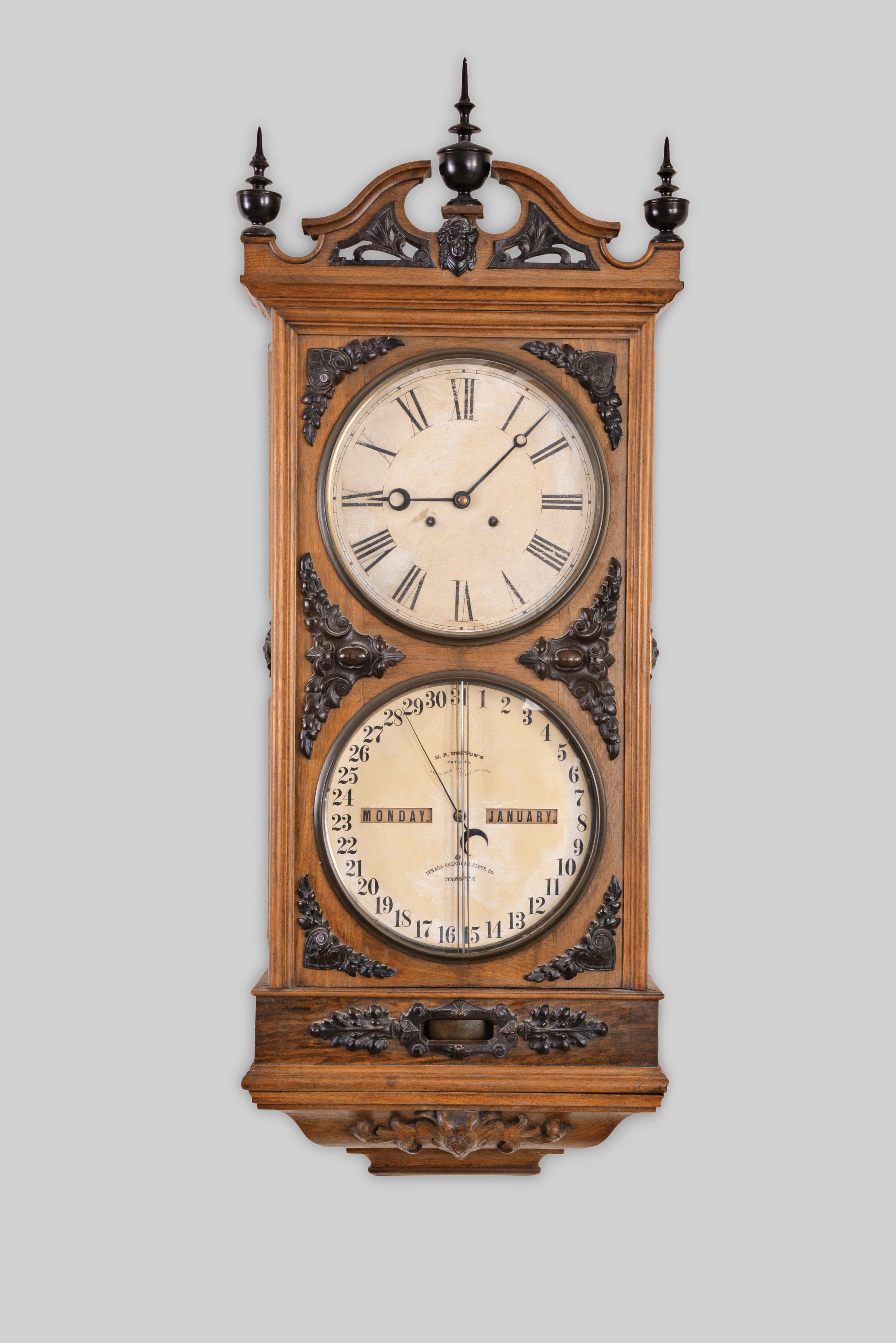 Ithaca Calendar Clock Co., Ithaca, New York, "Regulator No. 1" wall clock, 8 day weight driven Welch movement with perpetual calendar in a walnut case with three finals, ebonized composition ornaments, carved top and bottom and exposed pendulum rod forward of calendar dial with two papered metal dials. — Antique Clocks Price Guide