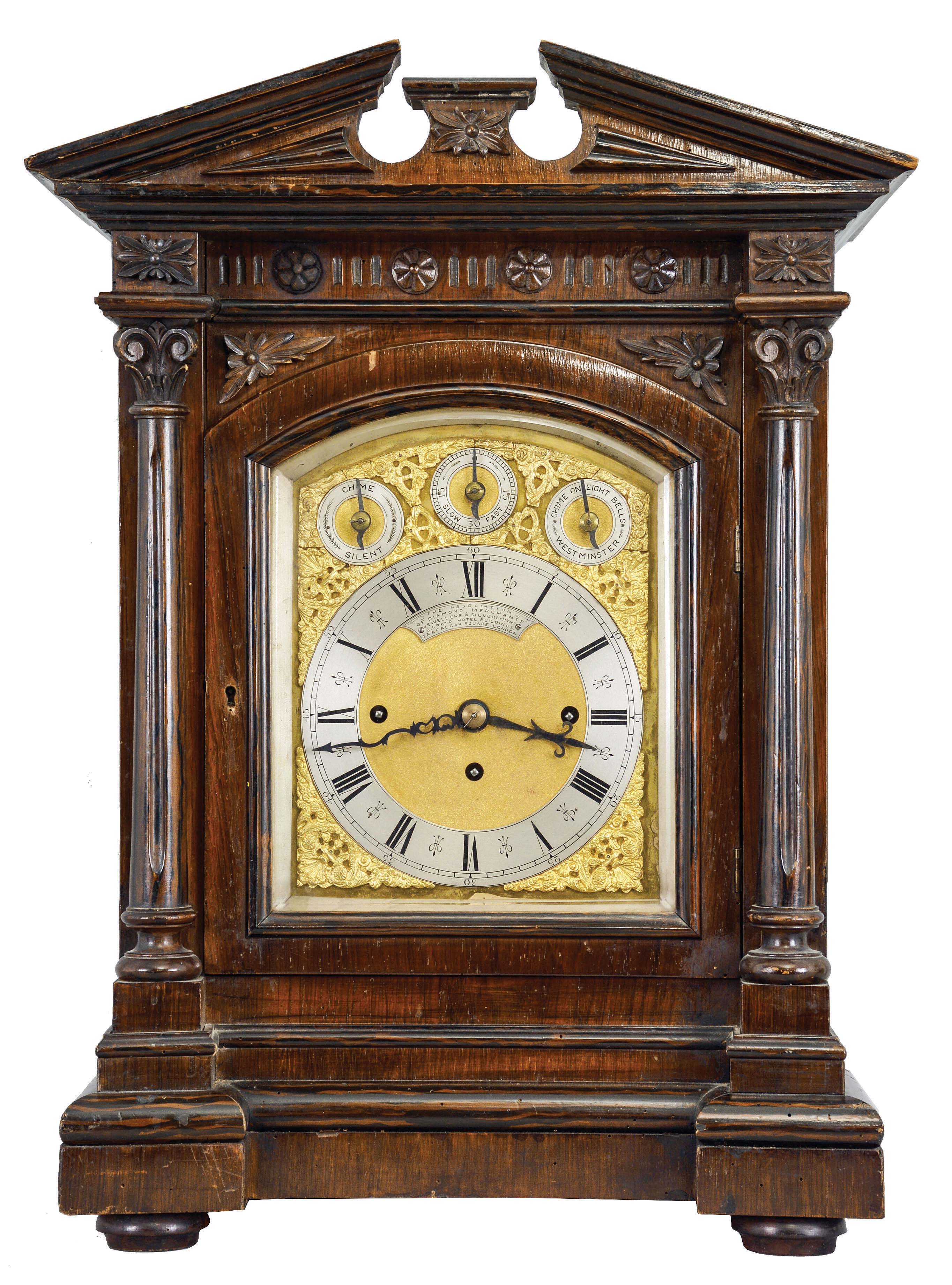 England, a monumental, quarter chiming table or bracket clock, the rosewood veneered and grain painted architectural case with molded base supporting a pair of columns with carved capitals below the carved entablature and broken pediment top, the sides with pierced, textile backed fret, composite dial with silvered roman numeral chapter ring, and subsidiary chime control and regulation at top, the dial also bearing an engraved plaque "The Association of Diamond Merchants, Jewellers, and Silversmiths, 6, Grand Hotel Buildings, Trafalgar Square, London", three train fusee movement chiming on eight bells or four gongs, and striking the hours on a larger, fifth gong — Antique Clocks Price Guide