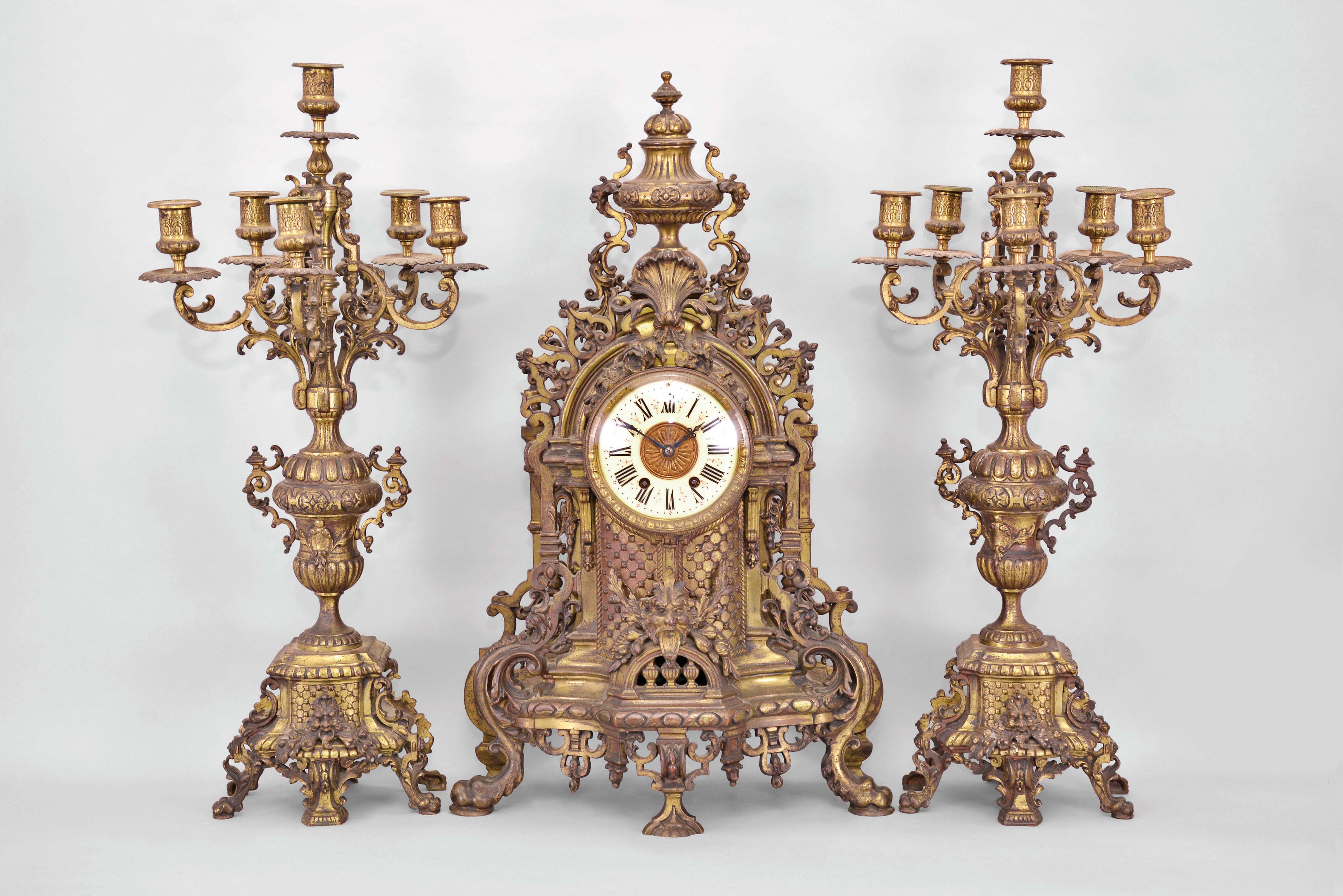 Jules-Edouard Dehon, Switzerland for Tiffany &amp; Co., New York, three piece mantel garniture set, 8 day, time and strike, spring driven movement in a elaborately sculpted cast brass Renaissance revival case featuring claw feet, foliate heads, floral motifs, peripheral fretwork, and lion masks in profile, cream porcelain dial with blued steel hands, all topped by a large urn finial. Two matching brass candelabra holding six candles each. — Antique Clocks Price Guide