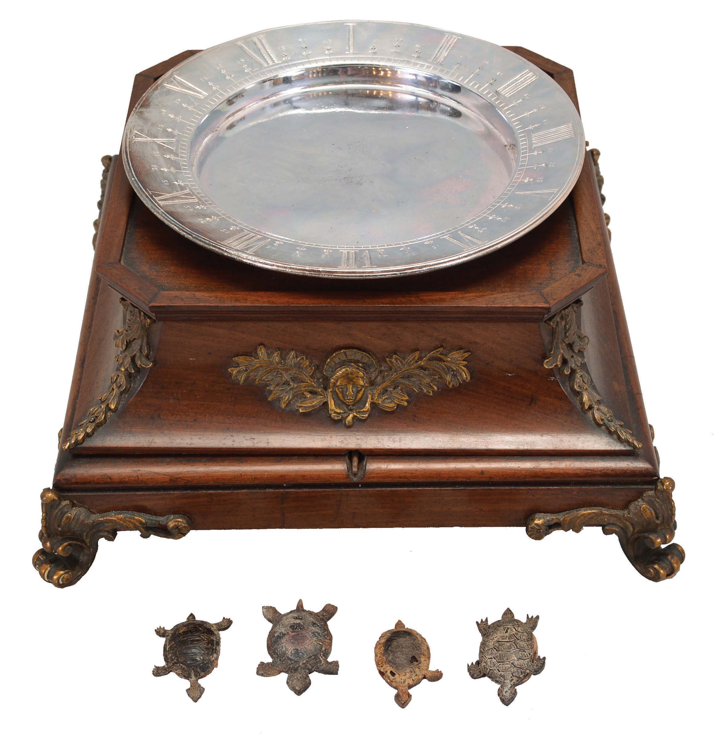 A. Cadot, Place Dauphine, Paris, a mahogany, gilt bronze mounted, floating turtle mystery clock, the case with square base on cast scroll feet, the upper portion with incurved sides with bronze mounts and molded top, supporting a water filled pewter bowl, the rim with engraved roman numerals, and arabic 15 minute markers, the turtle floating, and indicating the time, driven by a magnetic ring attached to the timepiece movement with lever platform concealed in the base, the back plate signed Samuel Marti, the front with Cadot's mark — Antique Clocks Price Guide
