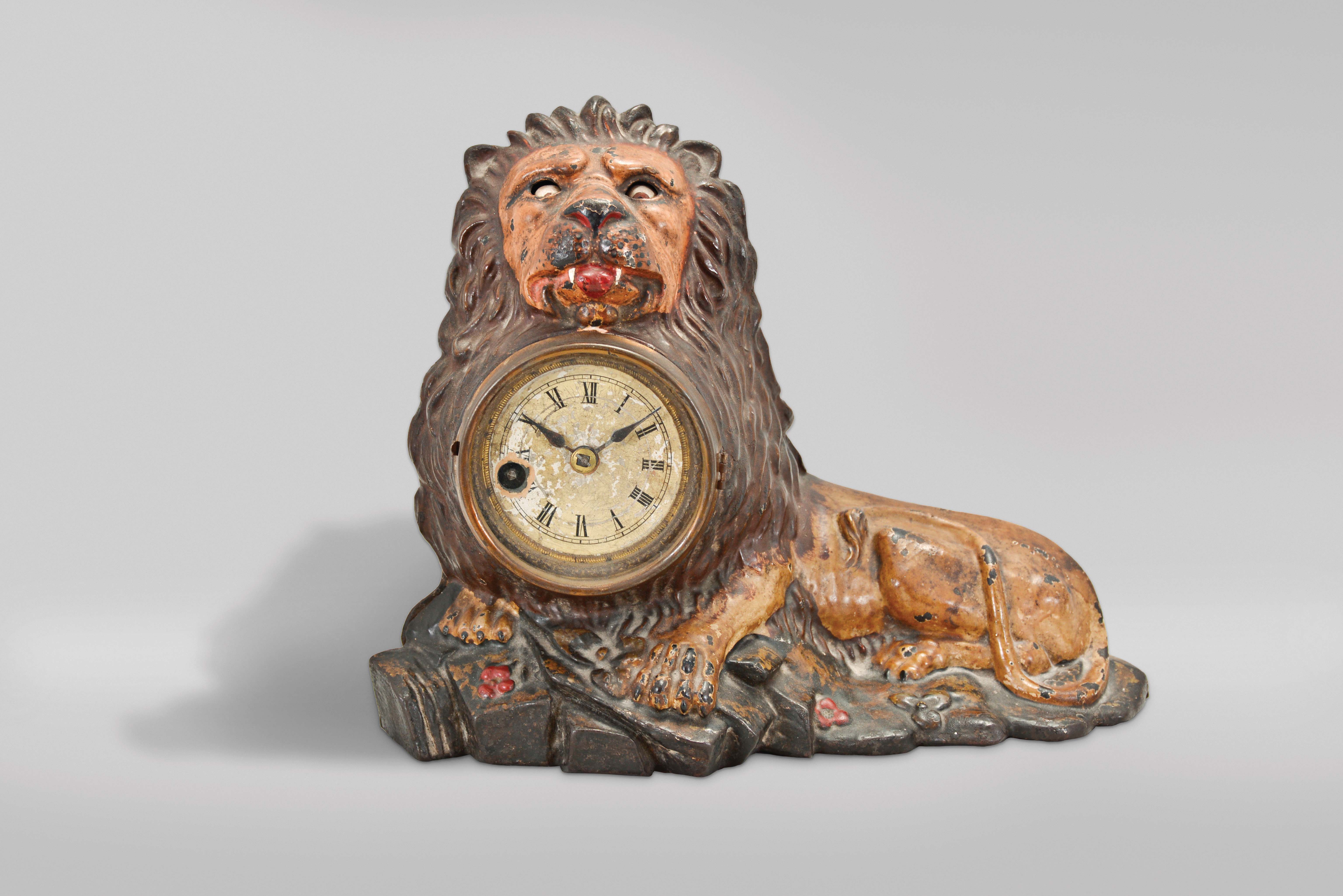 Bradley &amp; Hubbard, Meriden, Conn., "Lion" blinking eye shelf clock, the polychrome painted, cast iron beast recumbent in a rocky landscape and appearing to hold the bow of a giant pocket watch in his mouth, his eyes connected to a lever on the pallet arbor, the rocking arbor imparting an up and down motion to the eyes, roman numeral, varnished paper dial with blued steel spade hands, and 30 hour pendulum timepiece. — Antique Clocks Price Guide