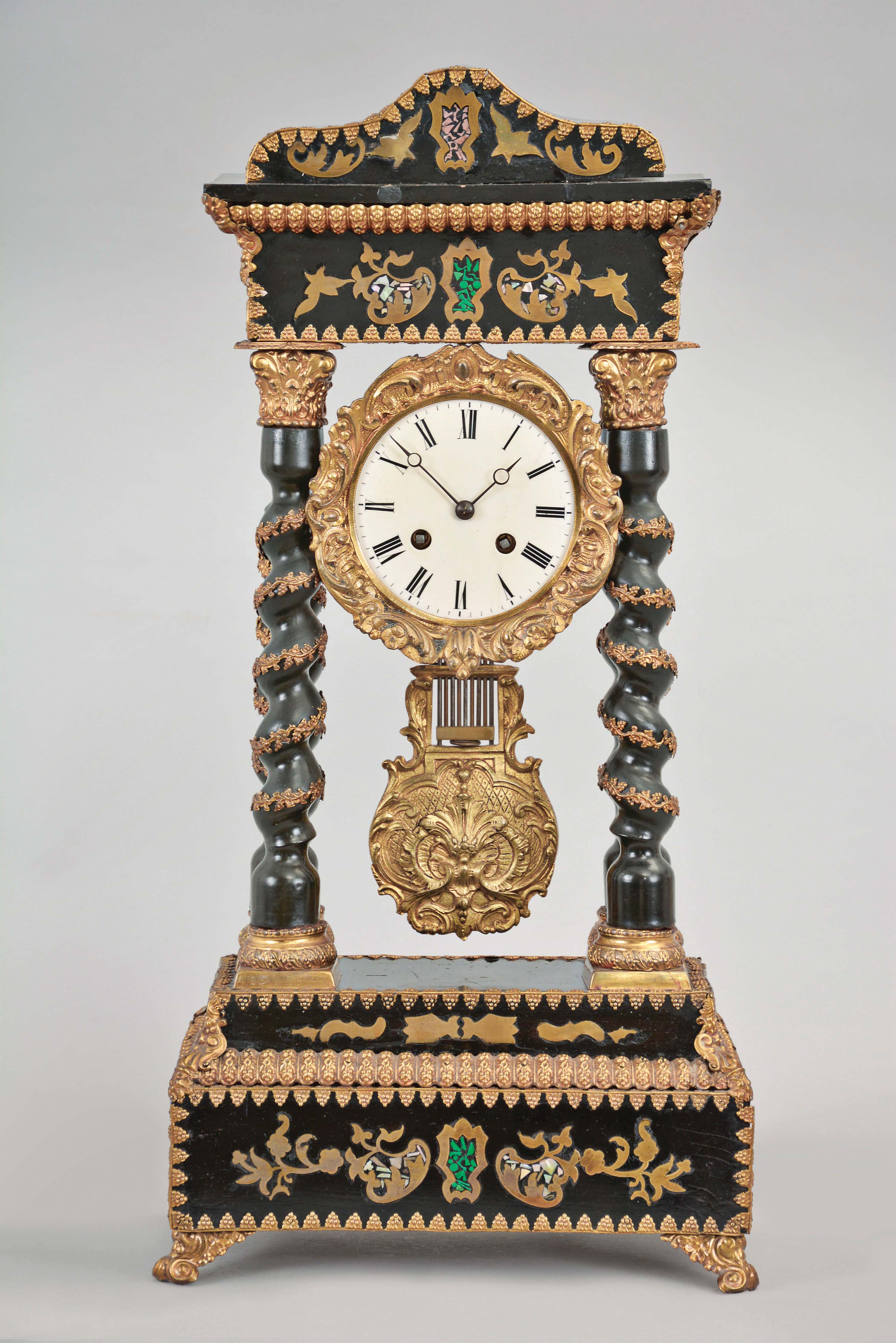 France, an indescribably outrageous mid 19th century portico clock, the stepped base, entablature and top inlaid with brass, mother of pearl, and green and pink dyed bone fragments, with stamped brass ornament and moldings trimming the ebonized case, twist turned columns with stamped capitals and bases, and trimmed with stamped brass vines, gilt, rococo revival style bezel with roman numeral white enamel dial, gilt gridiron pendulum with ornament matching the bezel, and 8 day, two train pendule de Paris movement striking on a bell — Antique Clocks Price Guide