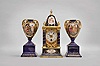 Austria, a Royal Vienna porcelain three piece garniture, the architectural clock case with cobalt blue ground, gilt, foliate ornament, and vignettes featuring putti in the clouds, gilt brass mid section surrounded by six porcelain columns, and containing the 8 day, timepiece movement, Arabic numeral white enamel dial, blued steel spade hands, and matching vasiform side pieces on square plinths, also in cobalt with gilt ornament, and each with courting scene in an oval reserve, signed "Hauffmann", all bases with beehive mark