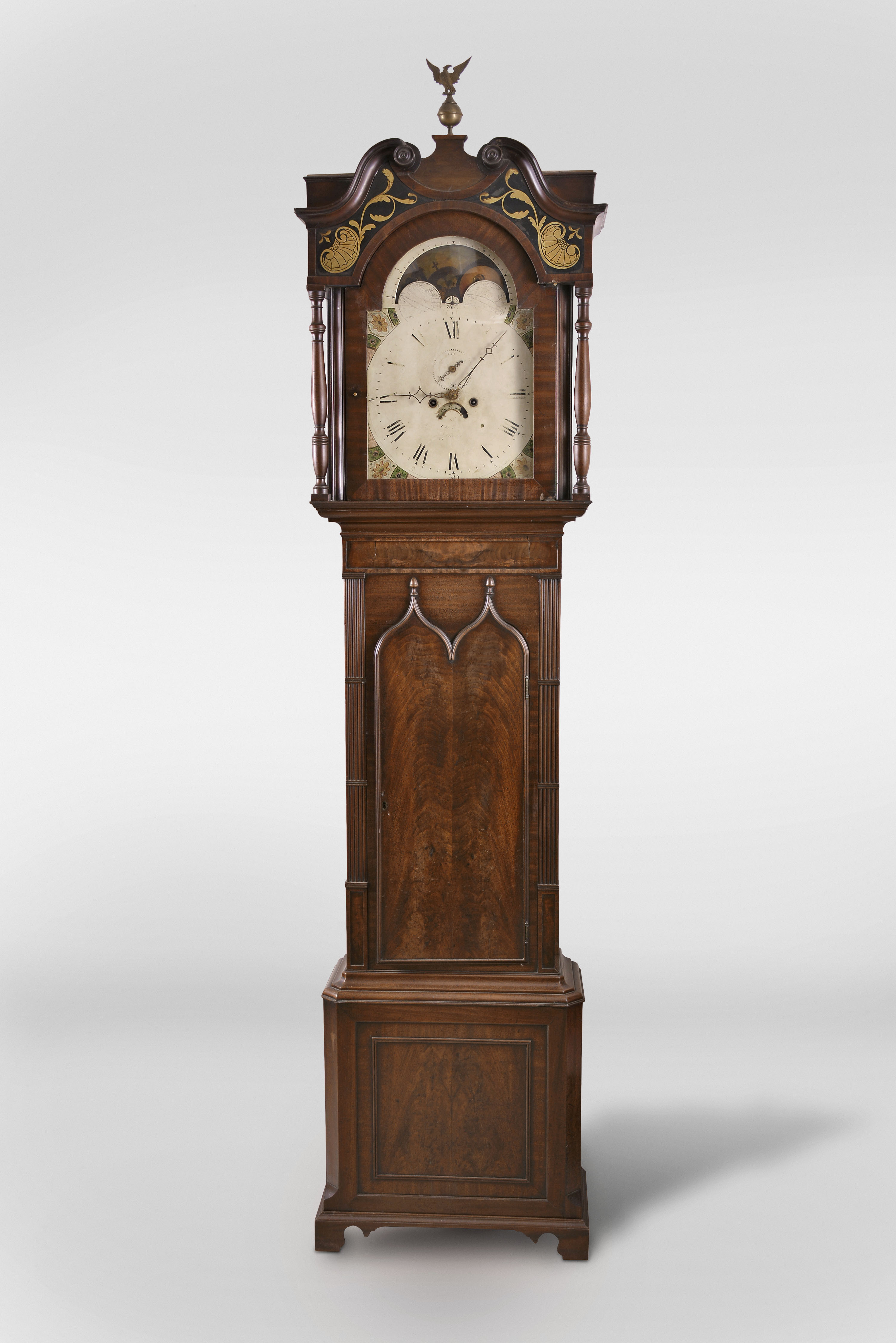 Charles Monks, Prescot, Lancashire, England, tall clock, 8 day, time and strike, weight driven movement in a Gothic revival mahogany case with reverse painted glass inserts above hood door, Gothic shaped trunk door over a base with framed panel on straight bracket feet. The painted metal dial has moon phase, seconds bit and calendar. — Antique Clocks Price Guide