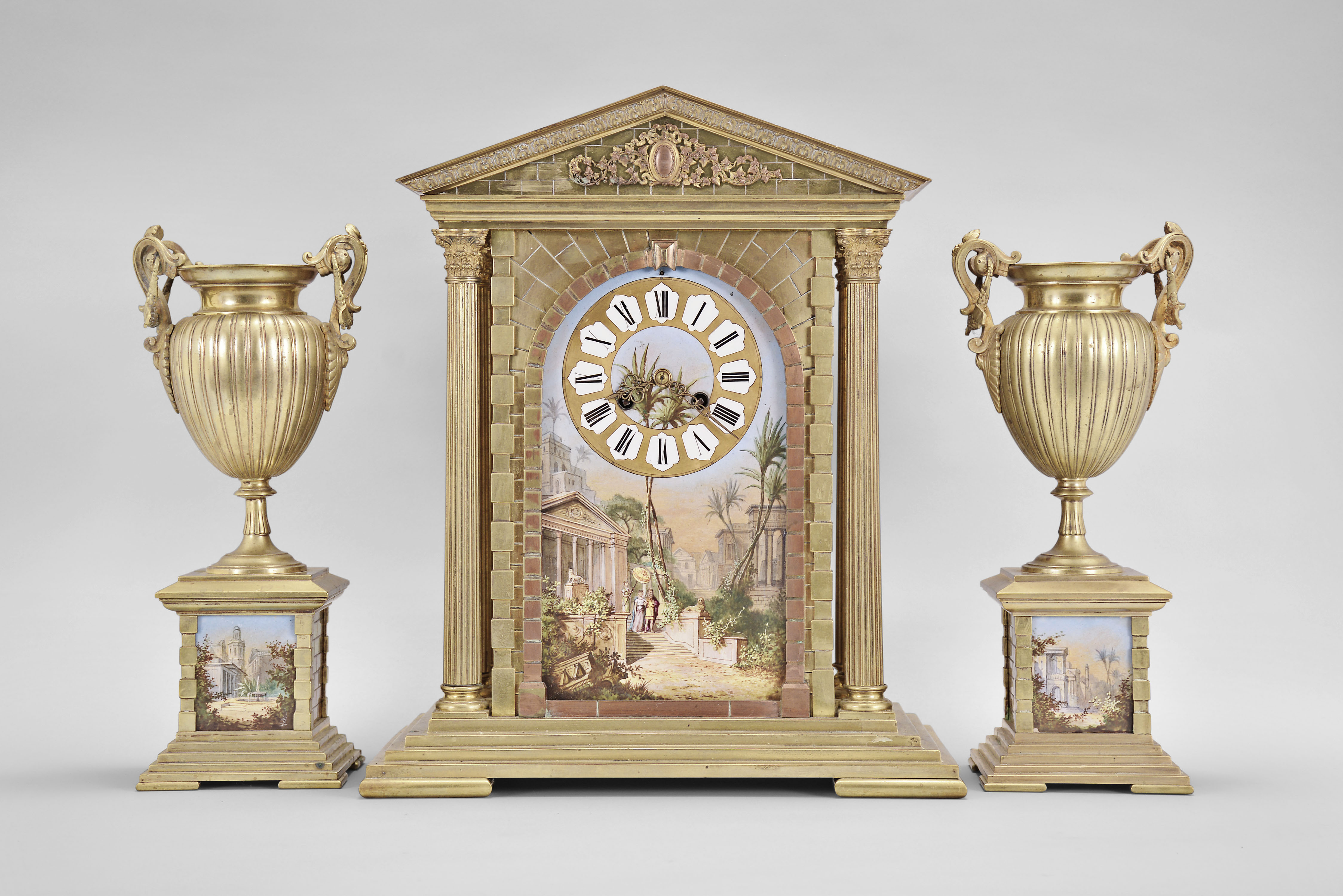 France, three piece mantel garniture set, 8 day, time and strike, spring driven pendule de Paris movement in a metal case of classic architectural form with stepped base, six full Corinthian columns flanking an ashlar facade with central arch, a painted ceramic panel within displaying a gilt chapter ring with Roman numerals, a scene of royals walking amidst an ancient city below the dial, all topped by a pediment with applied gilt ornament, two matching urns, both with painted ceramic panels. — Antique Clocks Price Guide