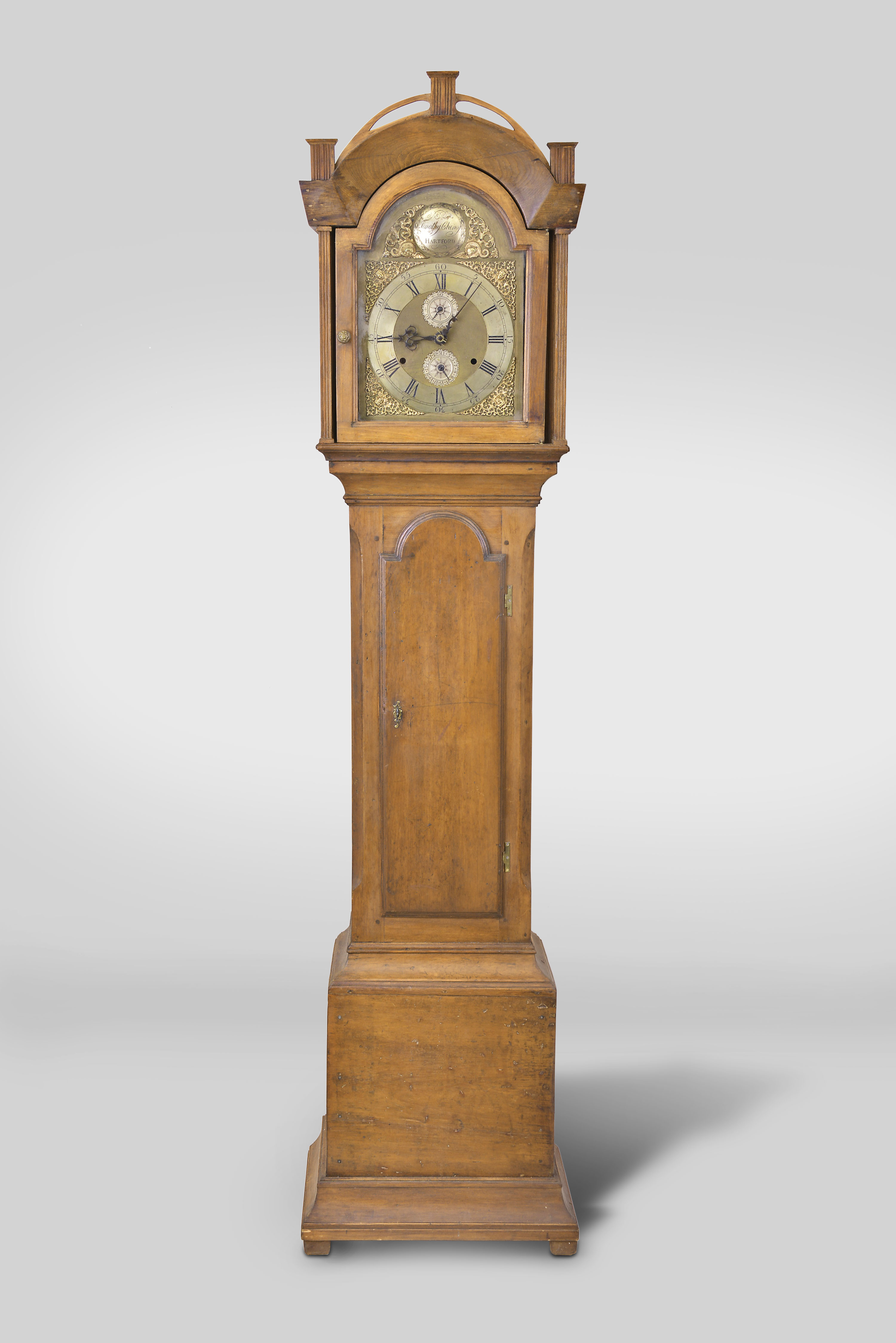 Timothy Cheney, Hartford, Conn., tall clock, 30 hour, time and strike, weight driven wooden movement in a pine case with arched top with reeded columns flanking hood door over a tombstone door on trunk resting on a cove molded and block feet. The brass dial is laid on wood and has a silvered chapter ring, calendar aperture, seconds bit and cast brass spandrels with Timothy Cheney signature in boss on arch. — Antique Clocks Price Guide