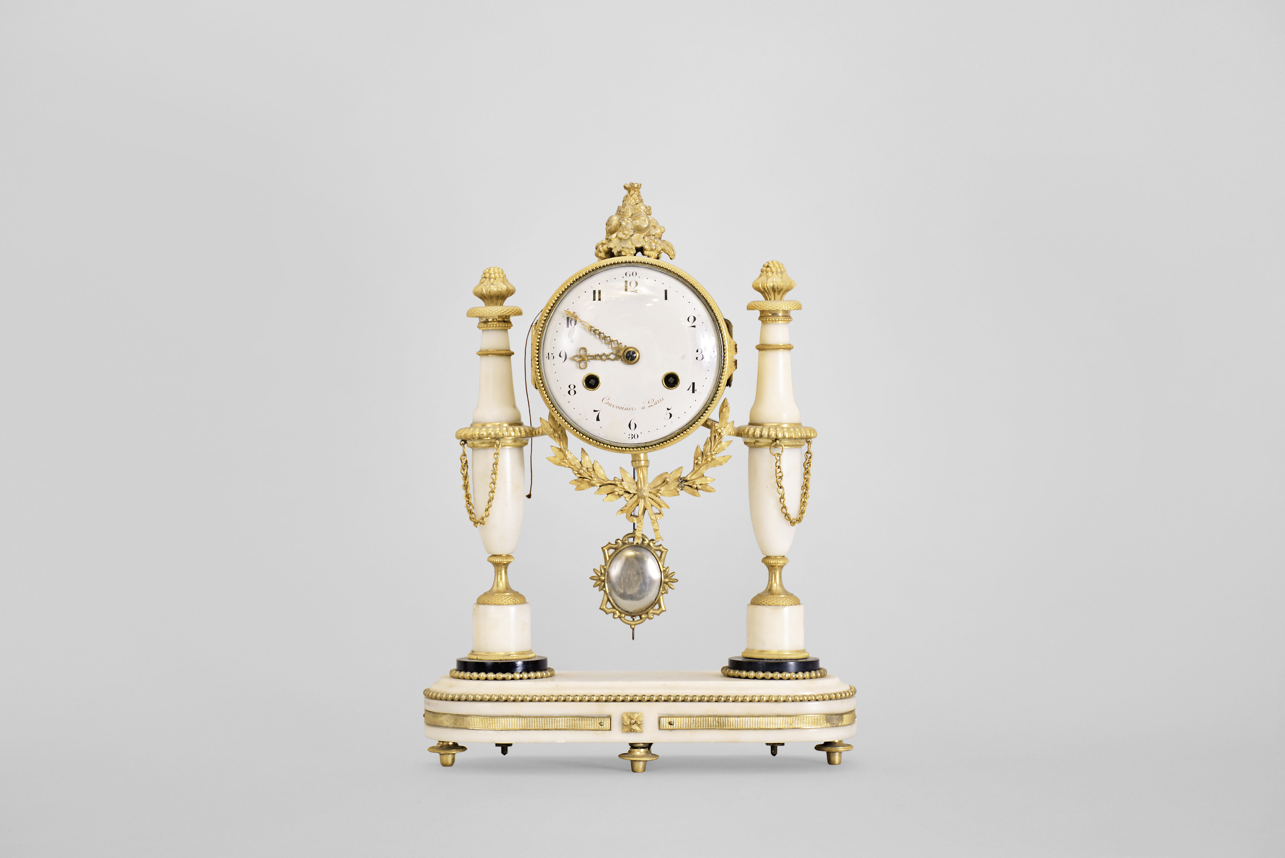 Courvoisier, a Paris, a late 18th century marble and gilt bronze mantel clock, the oblong base on toupie feet with inset, reeded mounts and central foliate ornament, and finished with a gilt bead molding, the gilt clock case with laurel and bowknot drop, and surmounted by a pyramid of fruit, the case supported bu two slender vasiform columns, with foliate and basket weave mounts, and gadrooned rings, arabic numeral white enamel dial, signed "Courvoisier a Paris", well executed pierced and engraved gilt hands, 8 day two train movement with crown wheel escapement, the Swiss style strike work mounted to the back plate, and with silk thread suspension, repeating the last hour on demand — Antique Clocks Price Guide