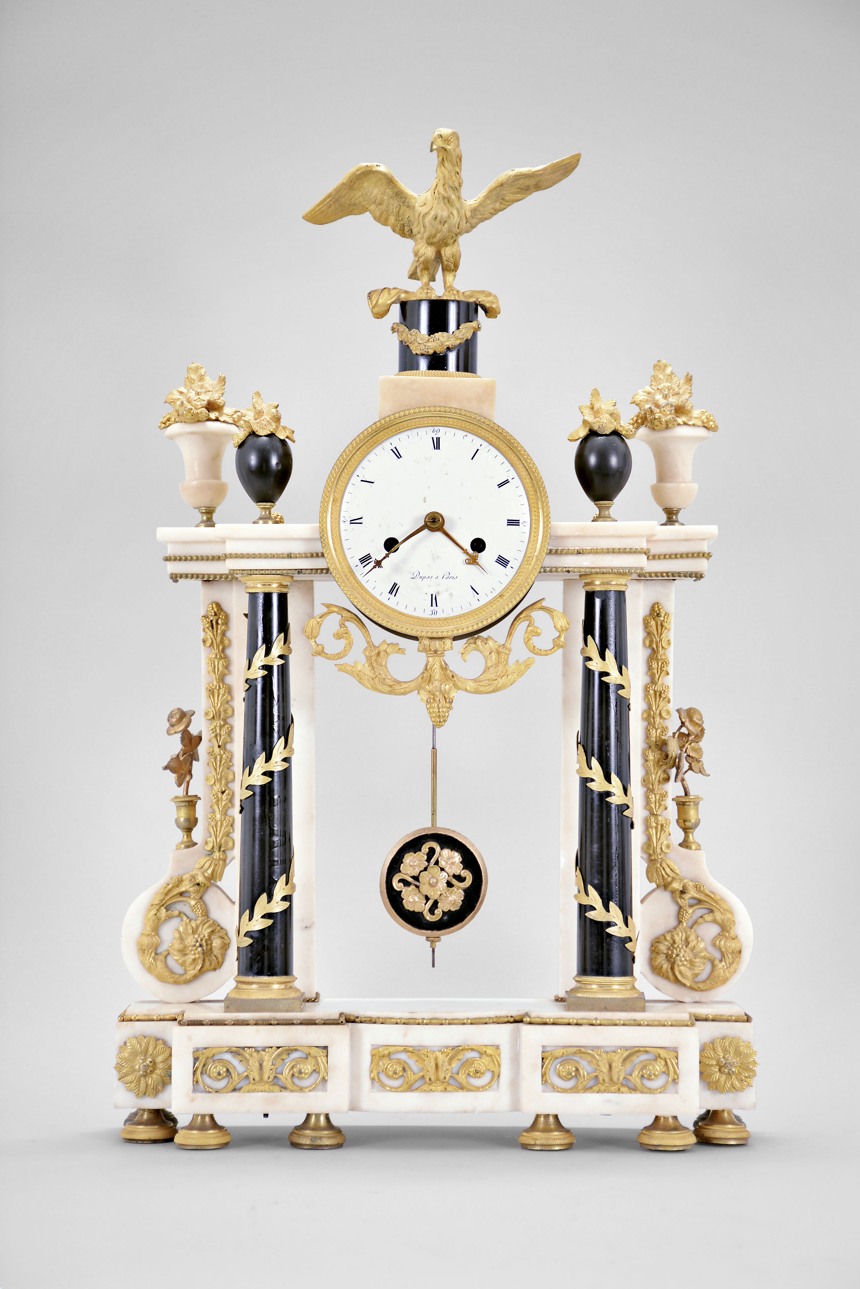 Dupas, a Paris, a late 18th century marble and gilt bronze mantel clock, the base on eight decorated feet, and with inset, scrolling foliate mounts, columns and pilasters supporting a stepped entablature with gilt bead molding, the columns in black marble with gilt capitals and bases, encircled with a spiral, leafy vine, the white pilasters with flanking, scroll buttress bases, mounted with a hanging garland, scrolling at the bottom, and terminating in a floral roundel, the clock case with scrolling foliate pendant, flanked by four urns with gilt flowers, and surmounted by an eagle with outstretched wings, and resting on a black pedestal with gilt festoon, roman numeral white enamel dial signed "Dupas a Paris", fancy gilt hands, 8- 15 day pendule de Paris movement with recoil escapement, silk thread suspension, and count wheel striking — Antique Clocks Price Guide