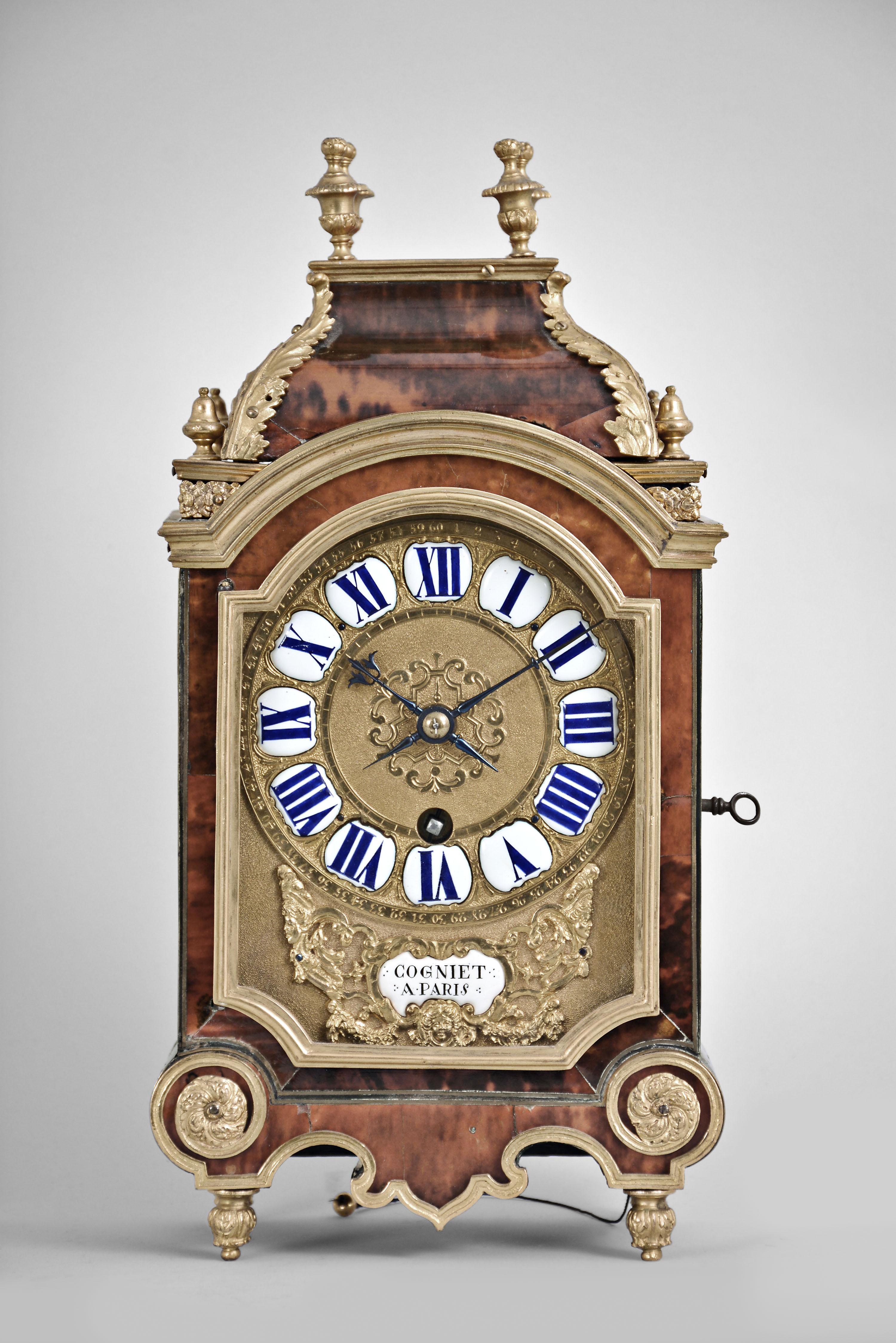 Cogniet, a Paris, an early 18th century brass and tortoise shell veneered, ormolu mounted pendule d' alcove, case with base on foliate ornamented feet, and having outcurved sides and scroll cut skirt, mounted with conforming gilt moldings terminating in foliate roundels, the gilt, cast door with incurved lower corners and arch top, and cast, molded cornice surmounted by a removable caddy top, with urn finials and foliate mounted corners, gilt brass dial with roman numeral cartouches above a cast ornament with masks, festoons, and central cartouche signed "Cogniet a Paris", fancy blued steel hands, signed brass movement with large barrel and crown wheel escapement, the striking activated by pulling a cord at the base, and sounding the last hour and quarter on two bells concealed beneath the top, with matching, incurved wall mounted bracket — Antique Clocks Price Guide